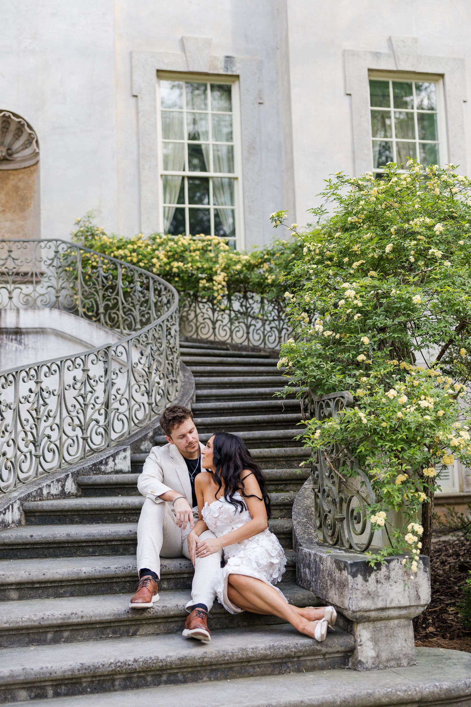 Elegant couple sitting on ornate stone steps of a classic building, surrounded by blooming yellow roses.