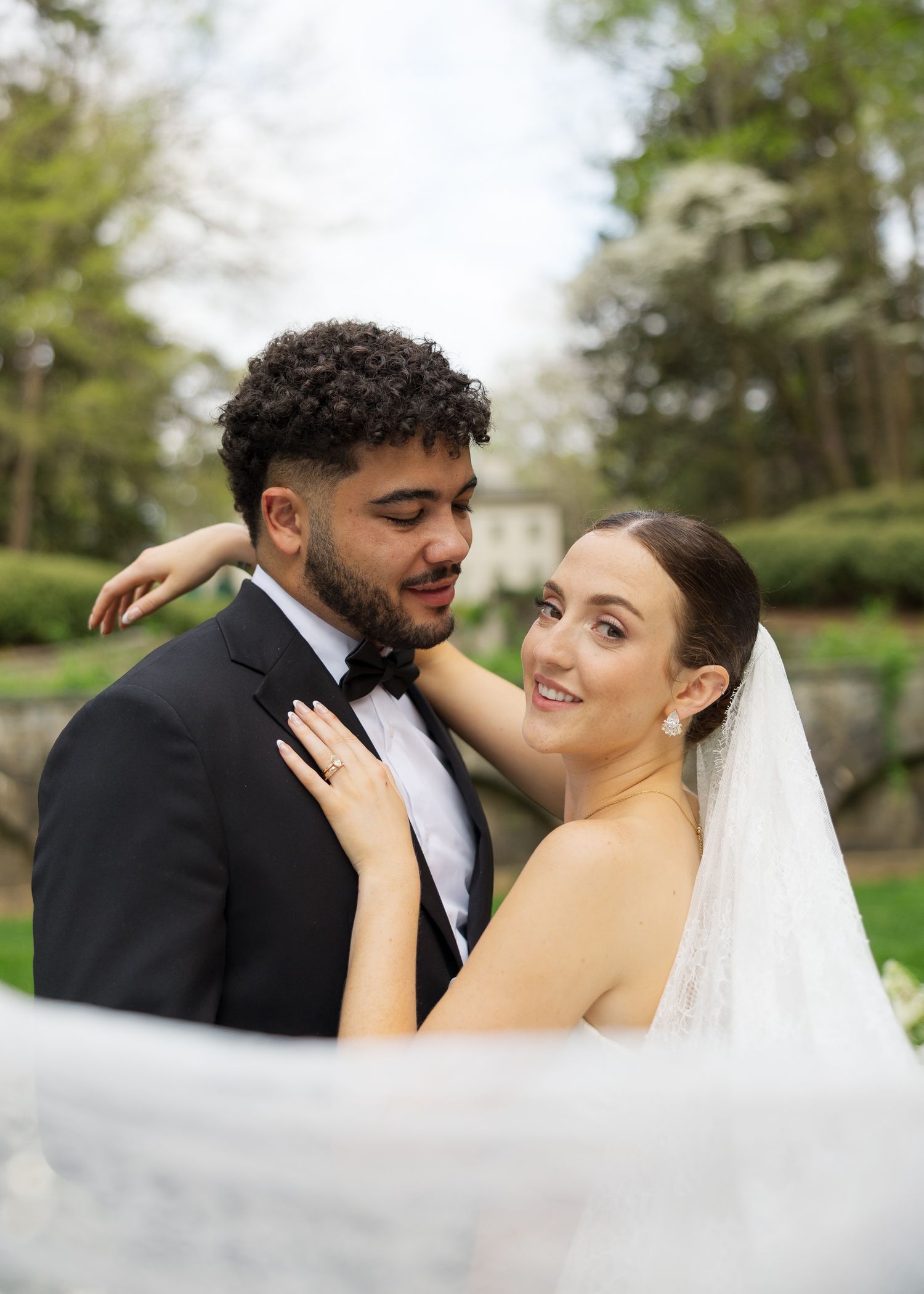 A bride and groom embrace outdoors in a lush garden, she wears a veil and he wears a black tuxedo.