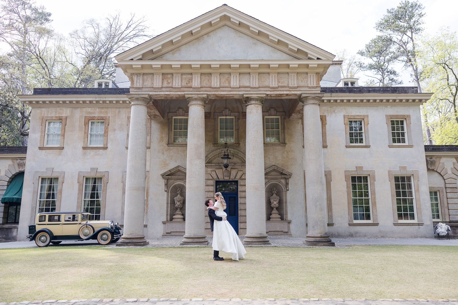 Bride and groom embrace before a grand neoclassical mansion with tall columns and a vintage car outside.
