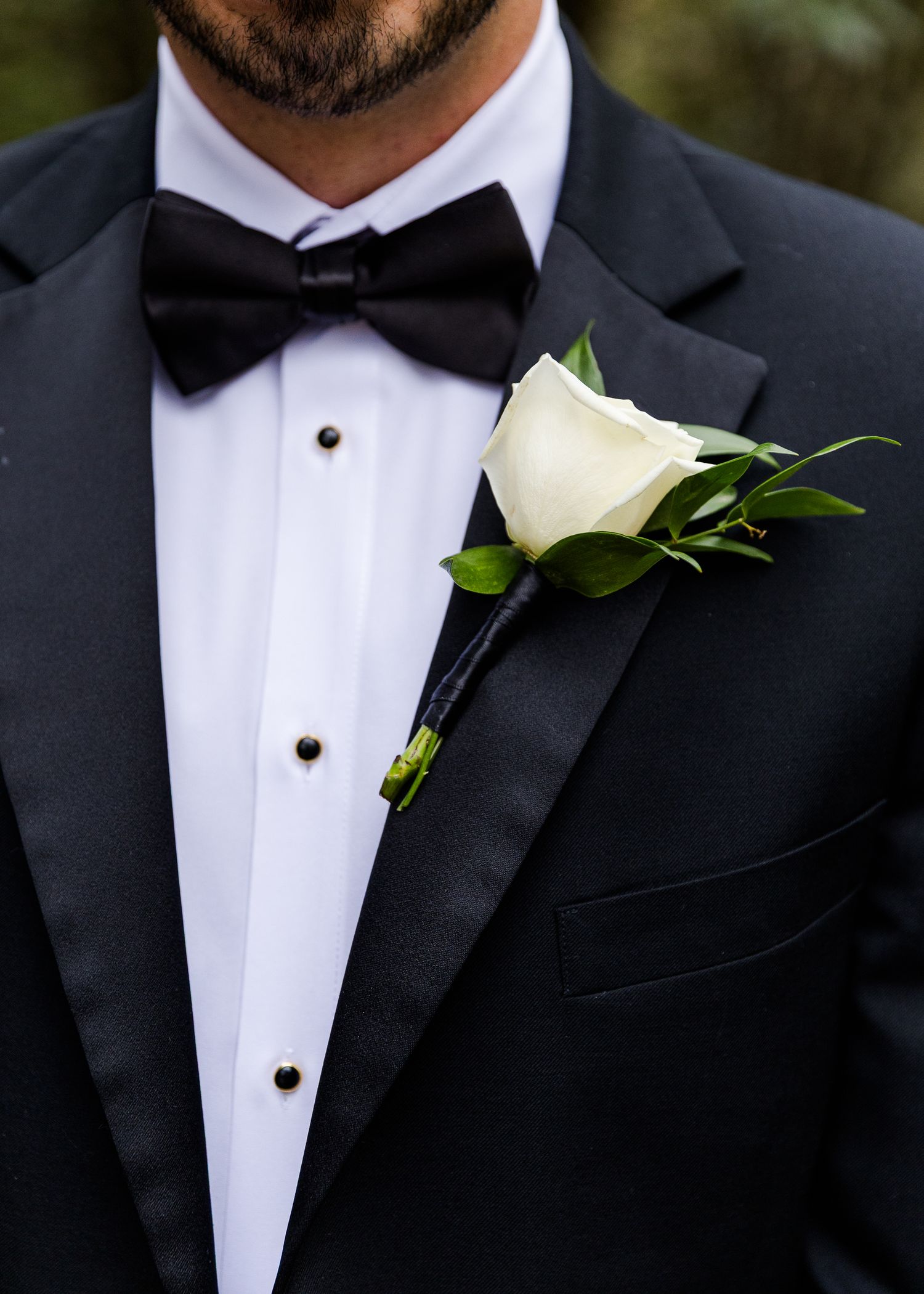 Groom in black tuxedo with bow tie and white rose boutonniere with greenery on lapel.