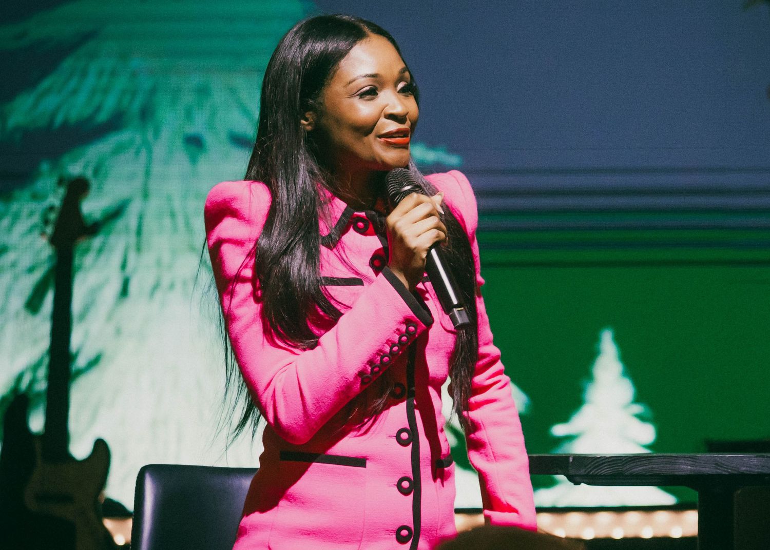 Performer in bright pink blazer singing on stage with green lighting effects.