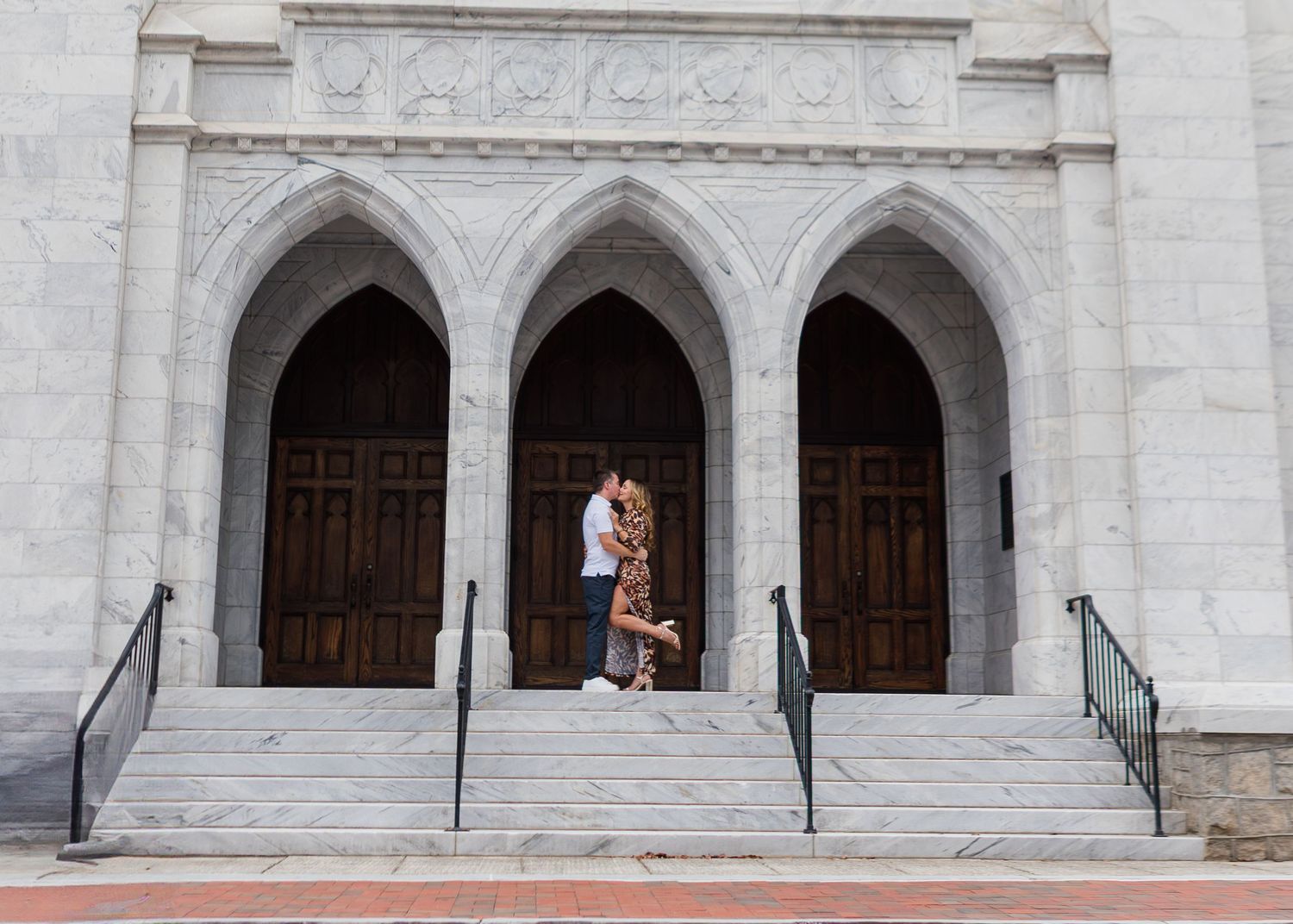 Couple embracing on steps of grand white stone church with arched wooden doors during engagement photo session.