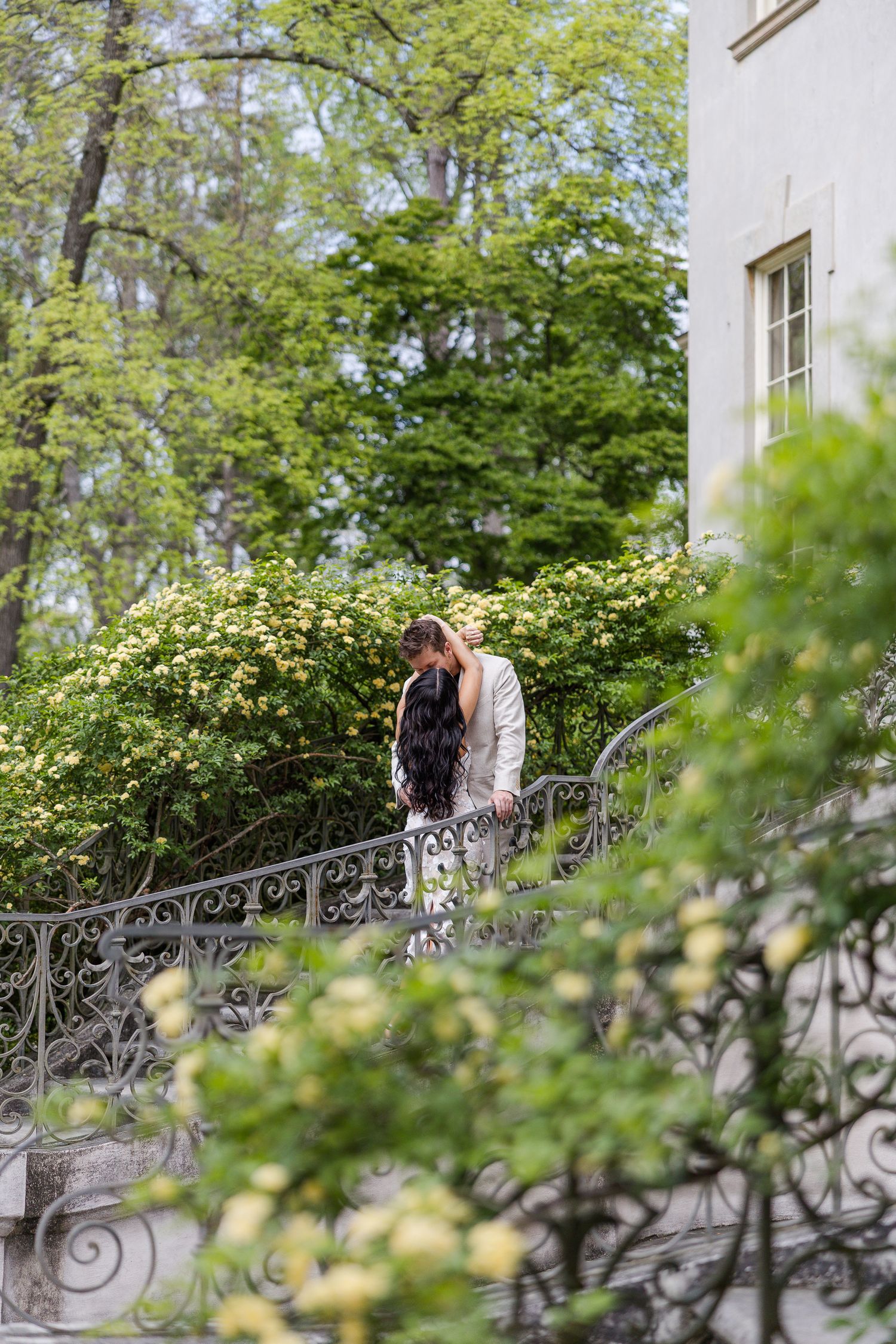 Couple shares romantic kiss on ornate iron staircase surrounded by blooming spring greenery.