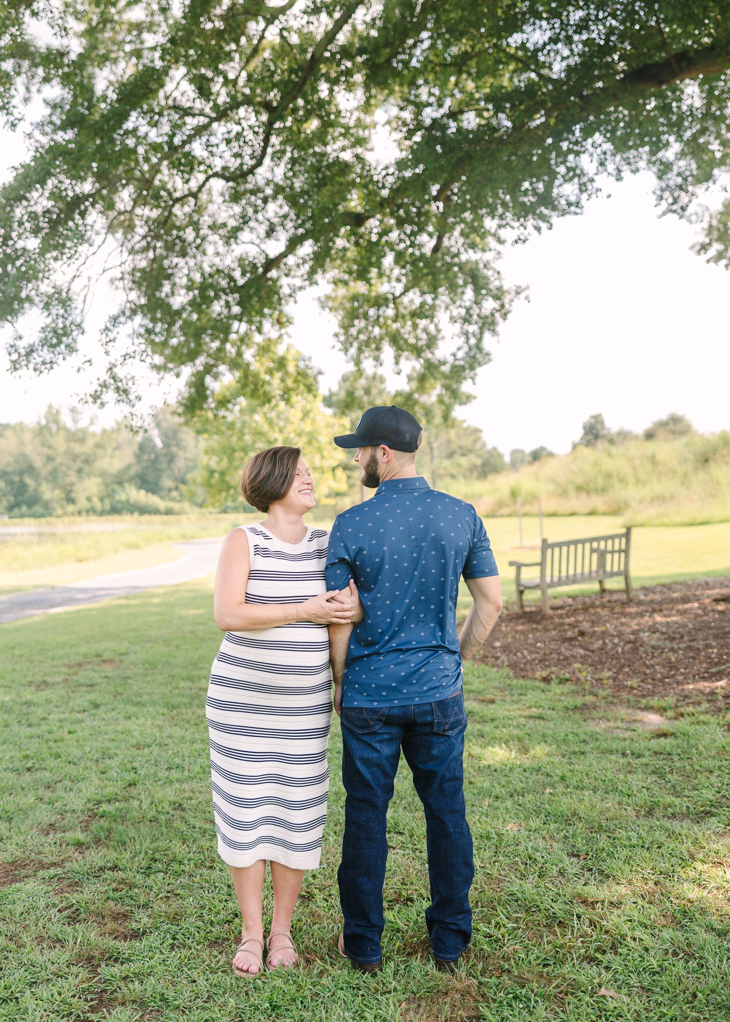 Couple standing together under a large tree in a sunny park, woman in striped dress, man in navy shirt and cap.