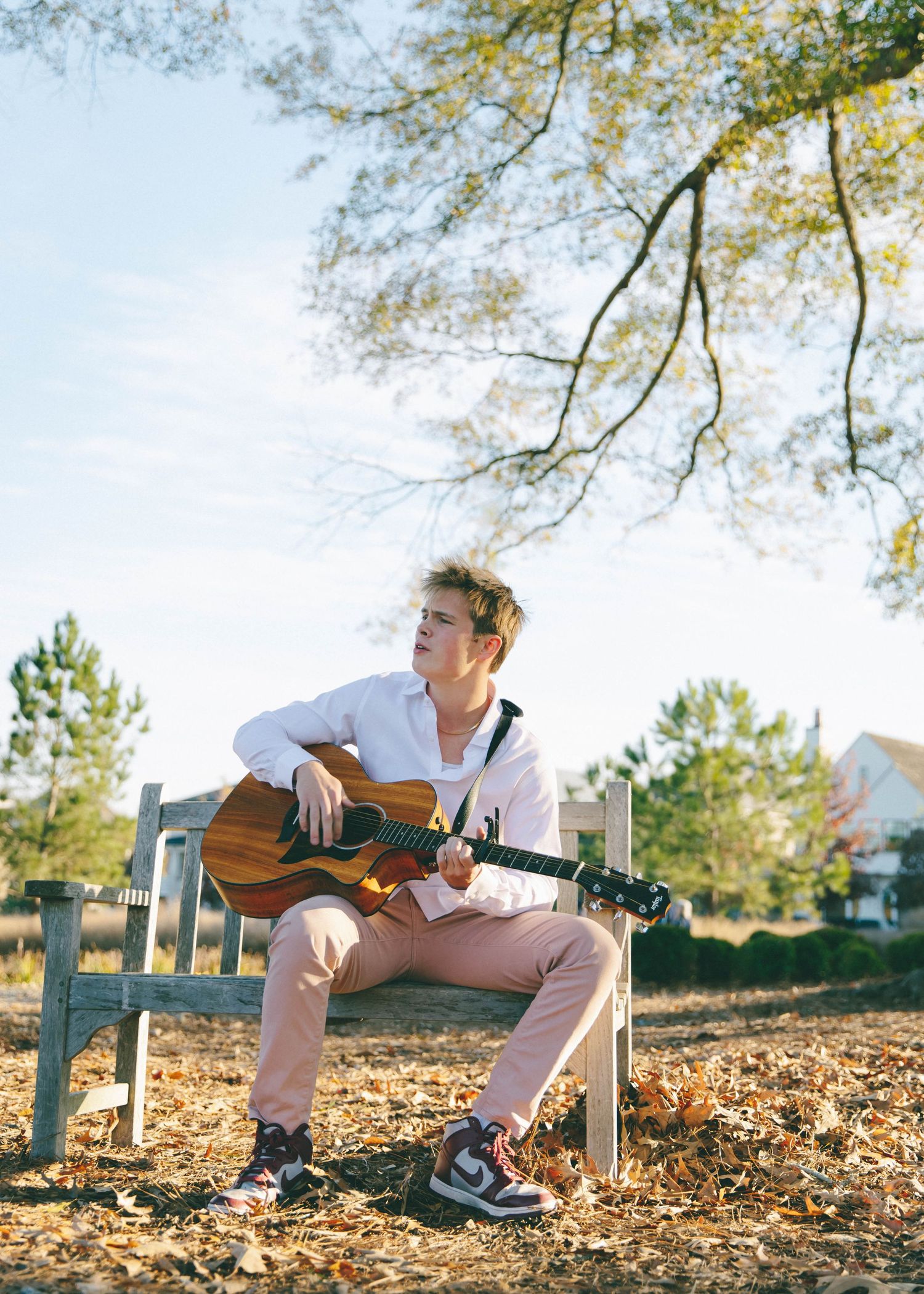 Person poses with guitar on wooden bench surrounded by fallen autumn leaves.