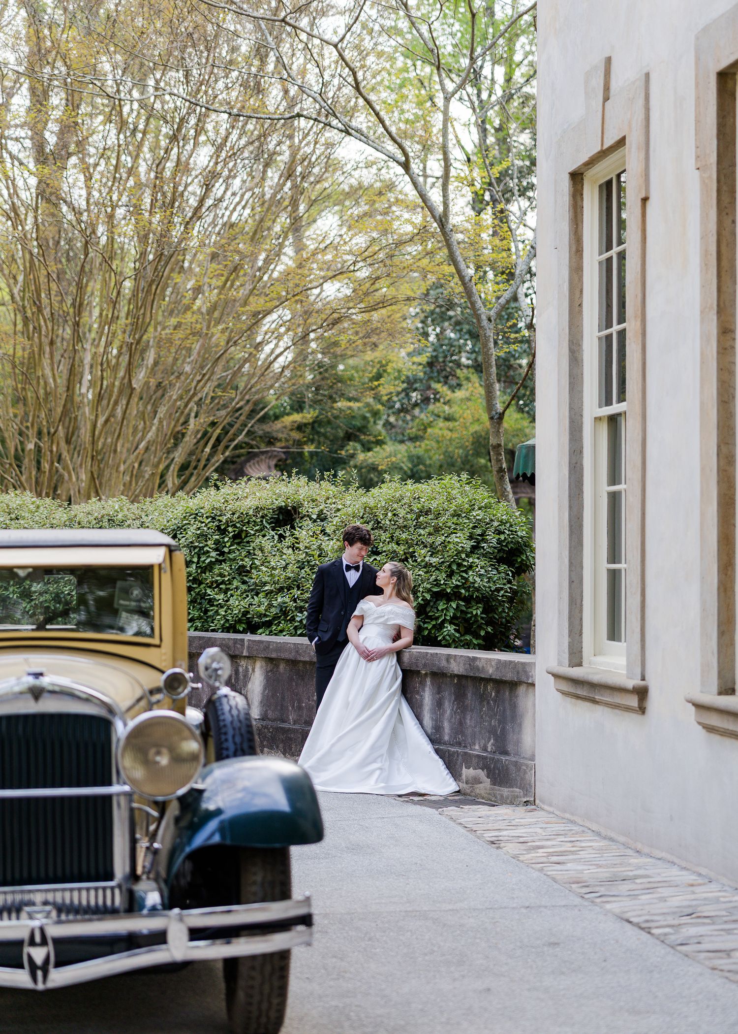 Bride and groom share a romantic moment beside a vintage car outside an elegant stone building.