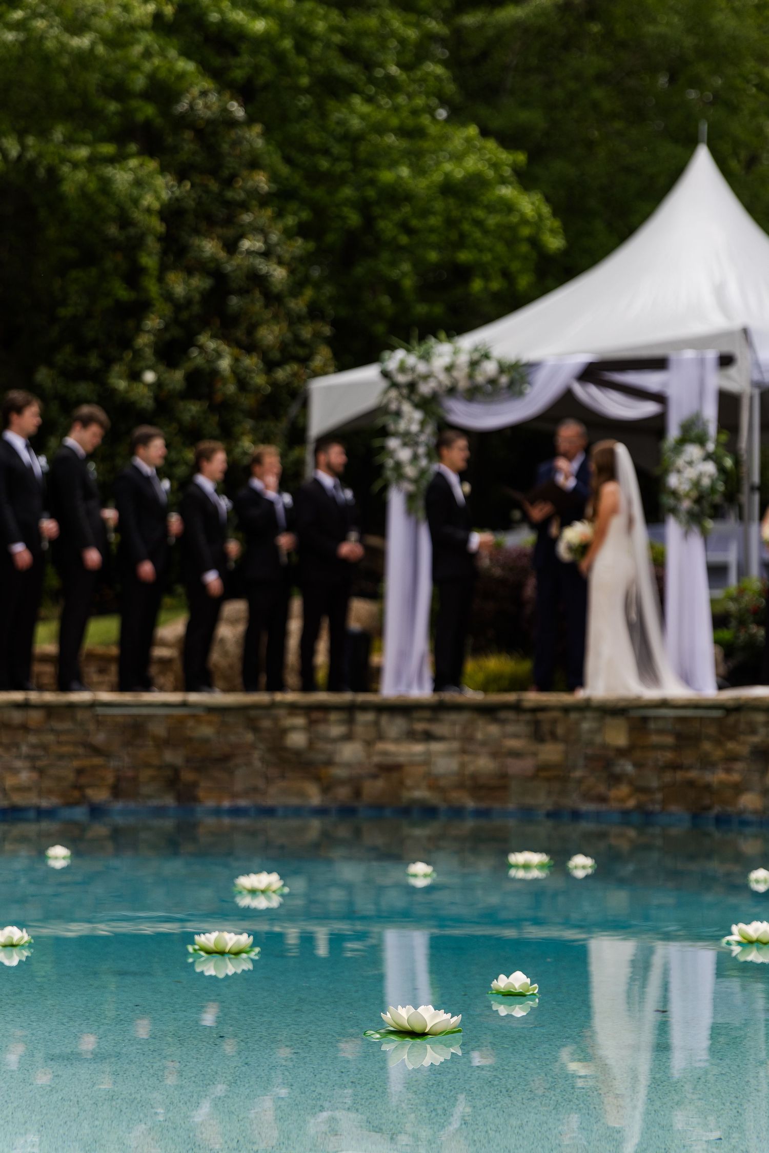 Outdoor wedding ceremony with groomsmen in black suits beside a floral arch, reflecting in a pool with floating white lotus flowers.