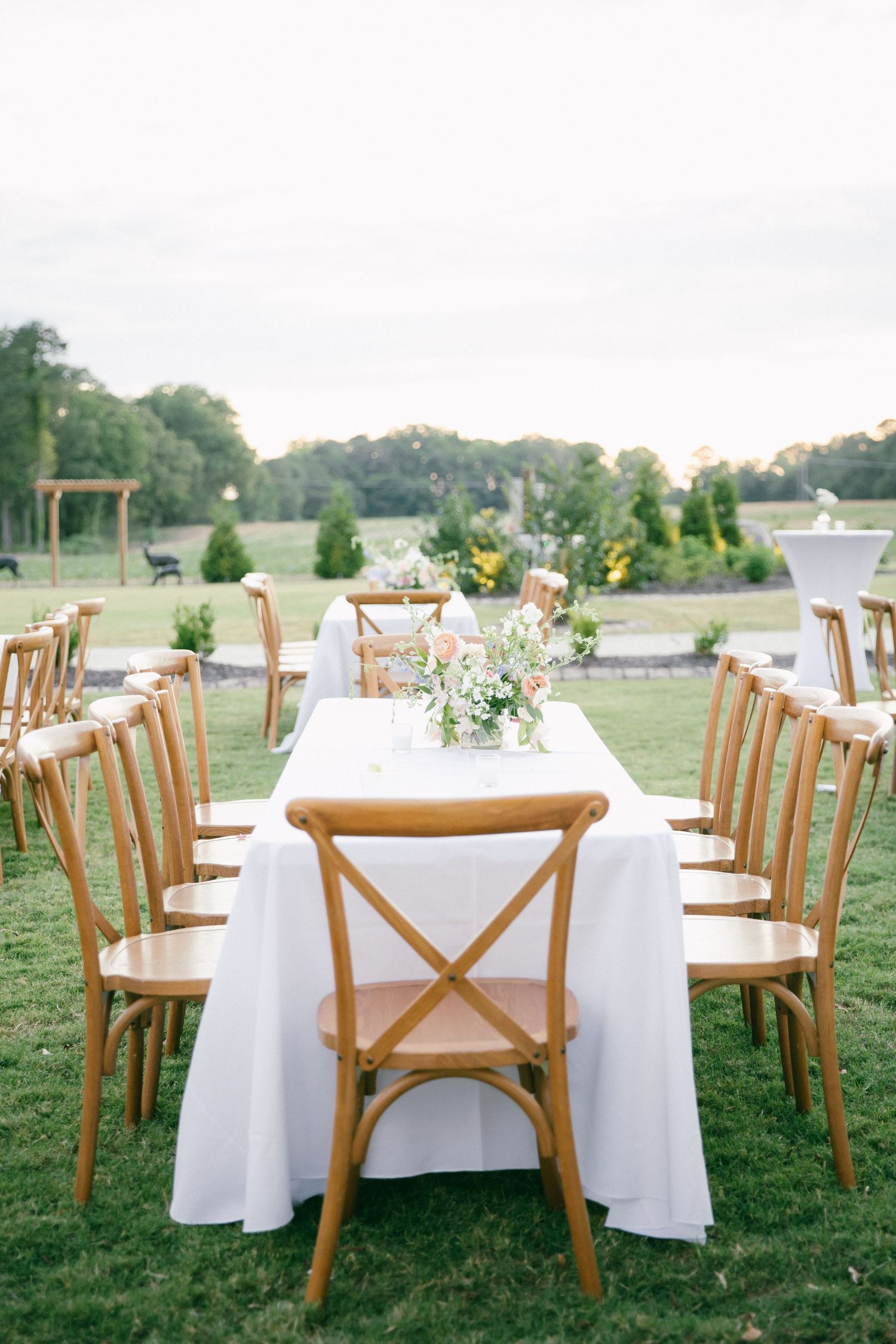 Outdoor wedding reception setup with wooden crossback chairs and white tablecloths.