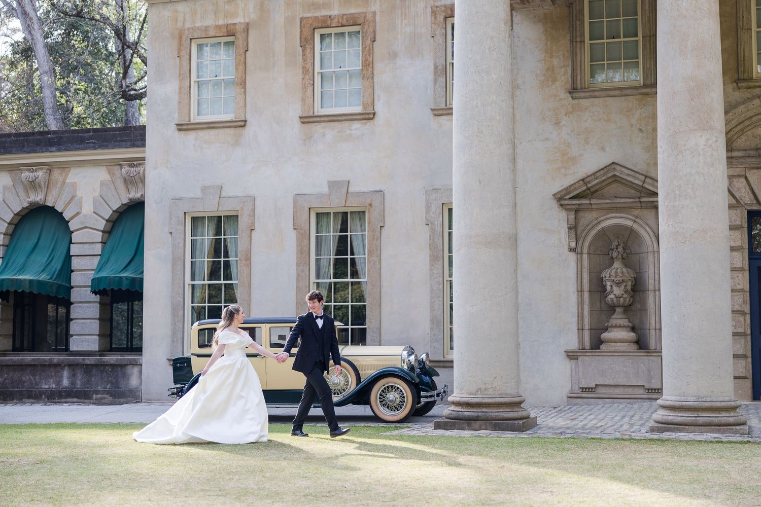 Bride and groom pose before a grand neoclassical mansion with a vintage cream car on a lush green lawn.