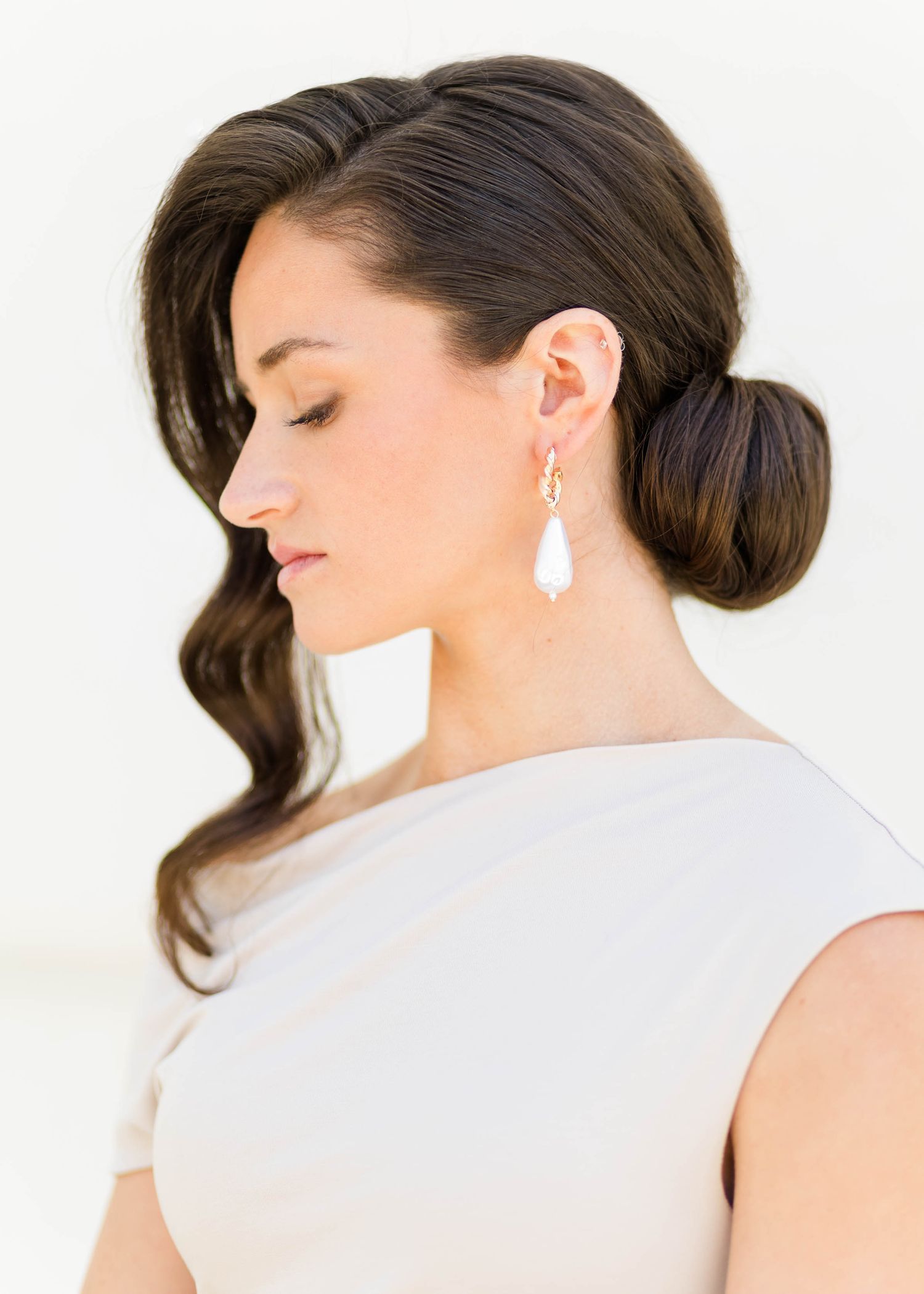 Elegant bridal updo with side-swept curl, pearl drop earrings, and sleeveless ivory dress on brunette model.