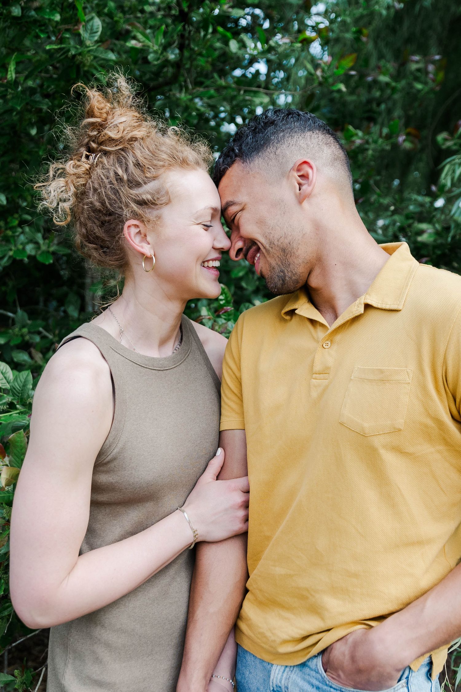 Happy couple touching foreheads and smiling together outdoors, surrounded by lush green foliage.