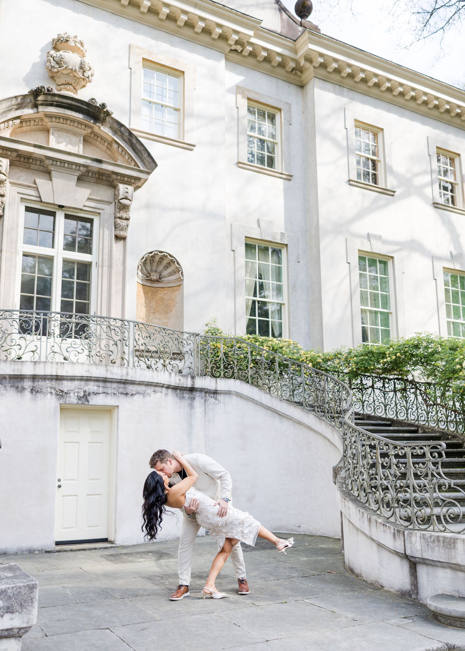 Couple sharing a romantic dip kiss in front of an elegant white Georgian mansion with ornate iron railings.