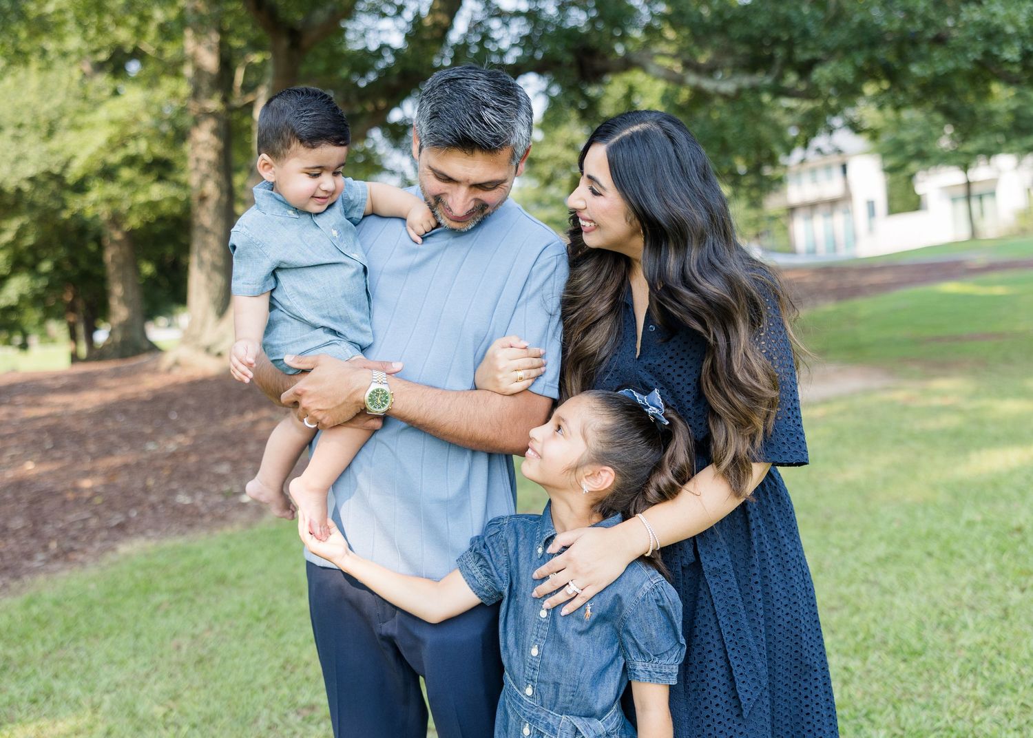 A happy family of four poses outdoors in a lush green park, parents embracing two young children in matching blue outfits.