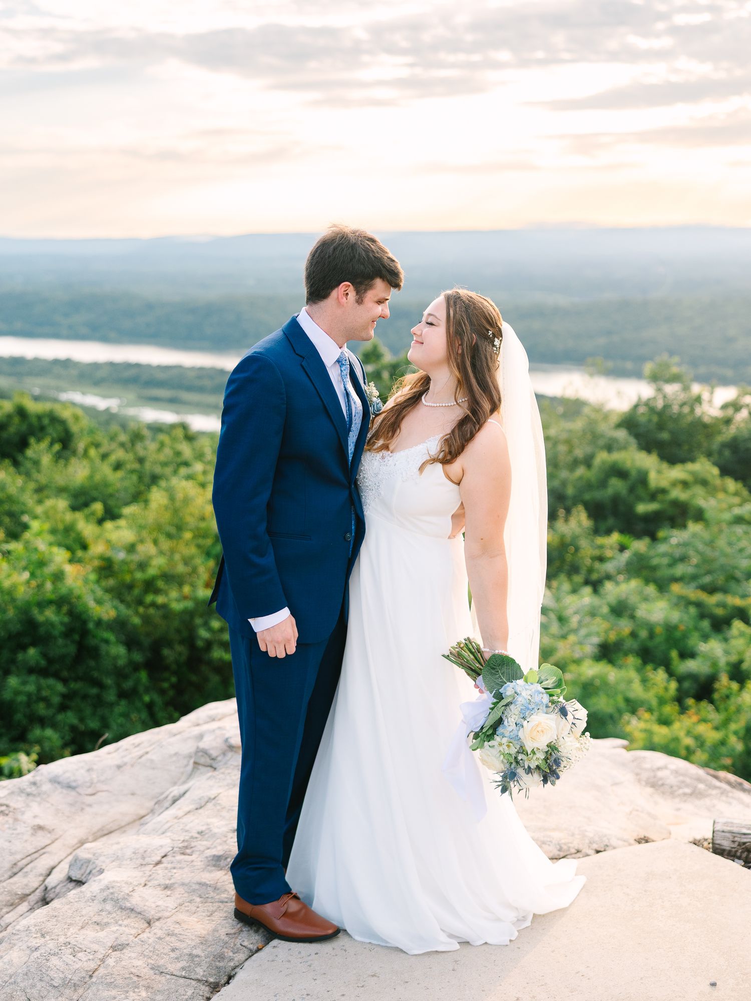 Wedding couple poses on rocky overlook with scenic mountain vista in background.
