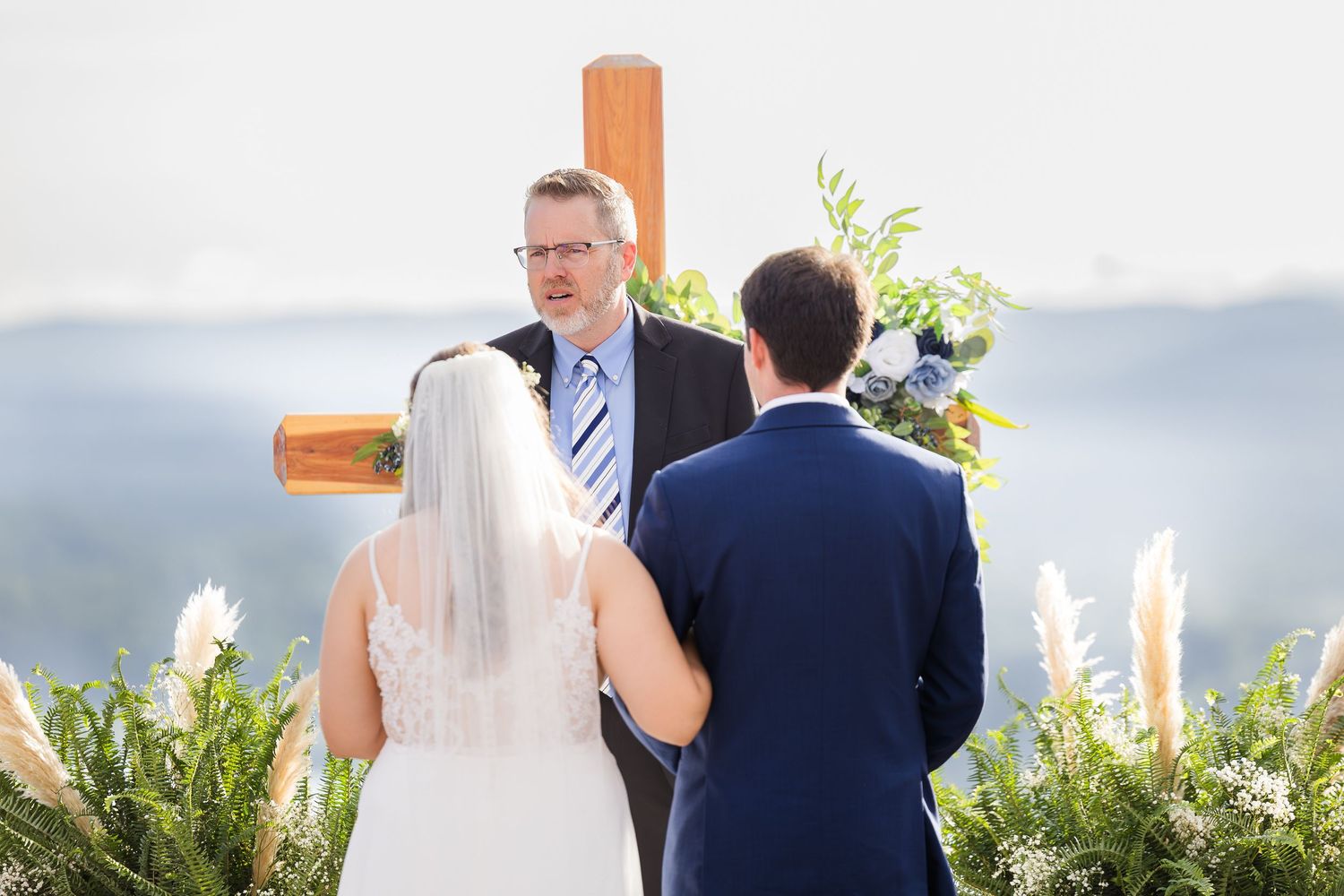 Bride and groom exchange vows before officiant at outdoor wedding ceremony with wooden cross and pampas grass.