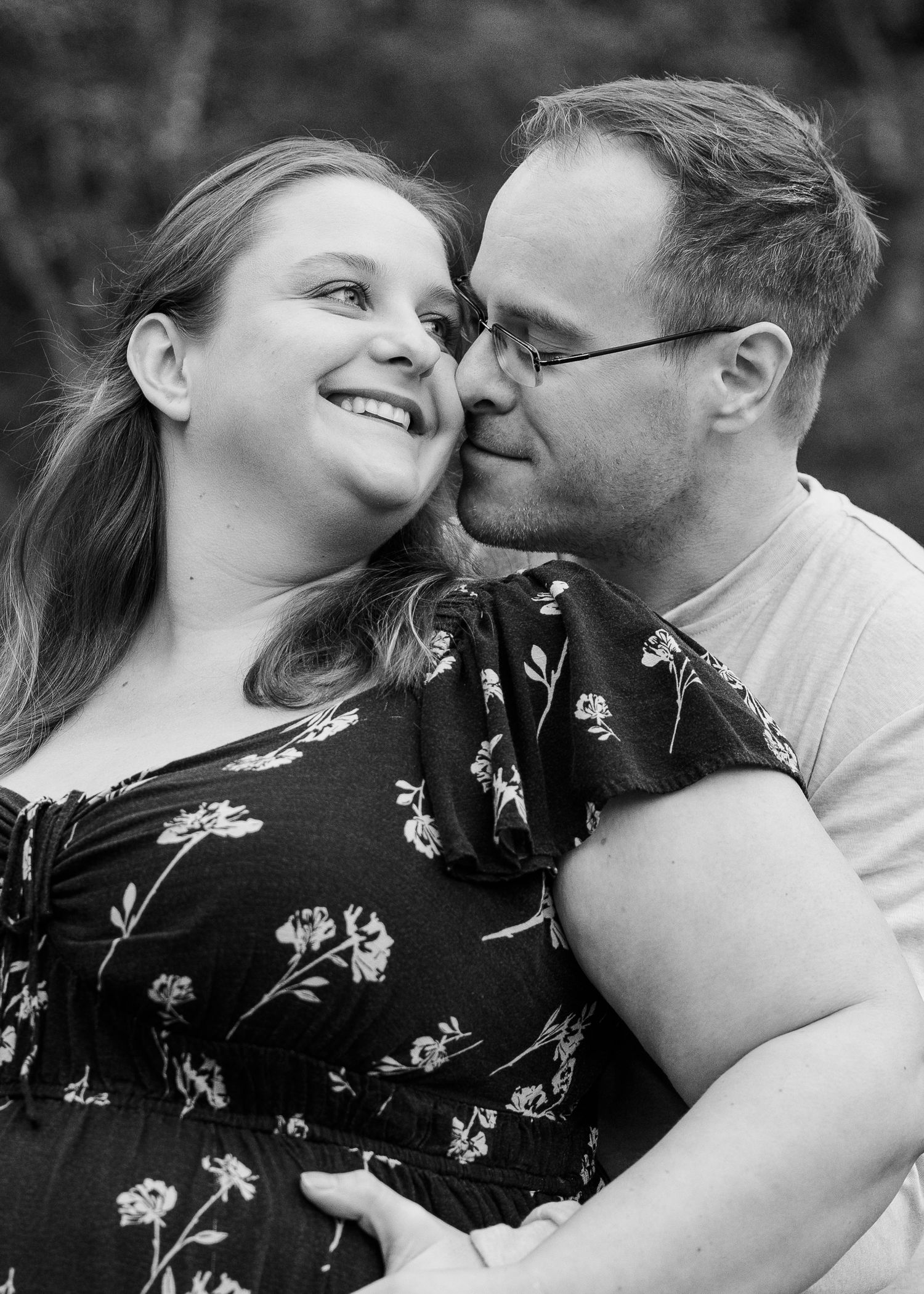 Black and white portrait of a smiling couple embracing outdoors, man kissing womans cheek affectionately.
