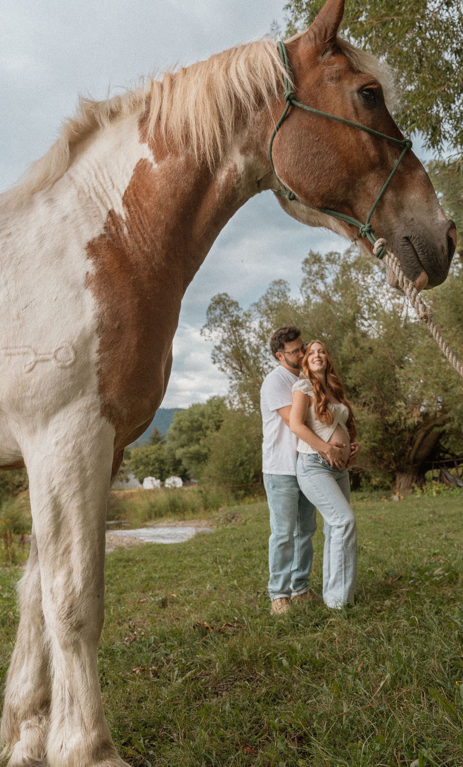 A couple embraces in a green field beside a brown and white paint horse wearing a rope halter.