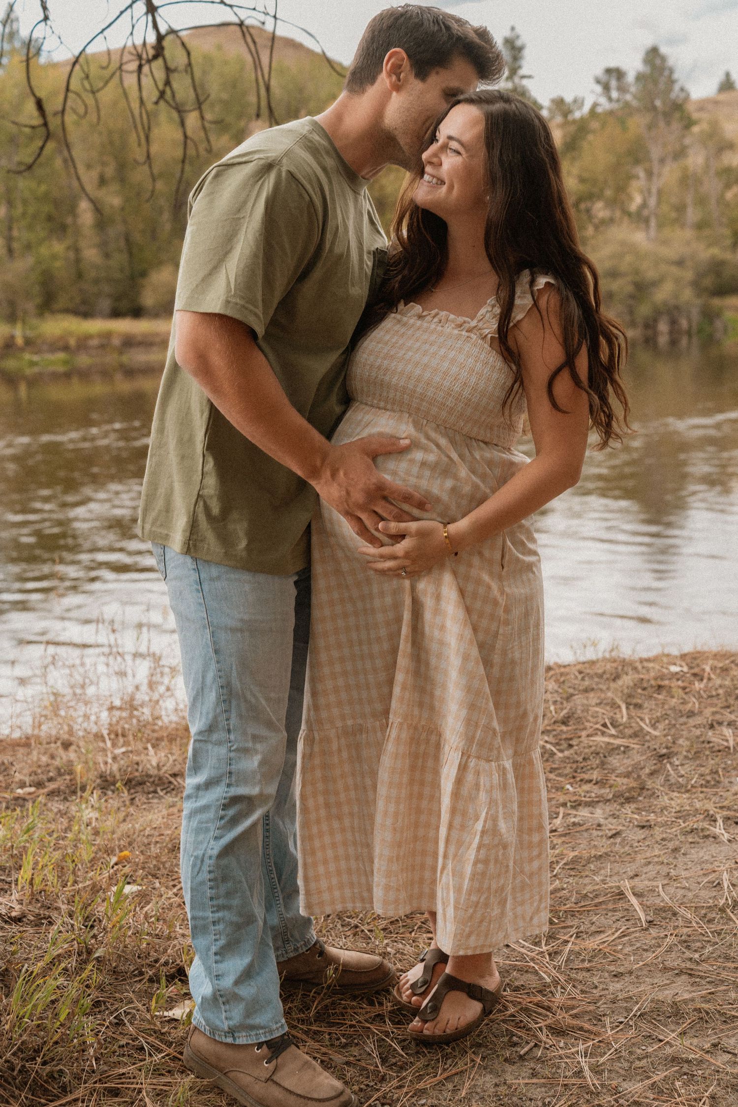 Couple sharing a tender moment by a lake, woman in gingham dress cradling baby bump during maternity photoshoot.