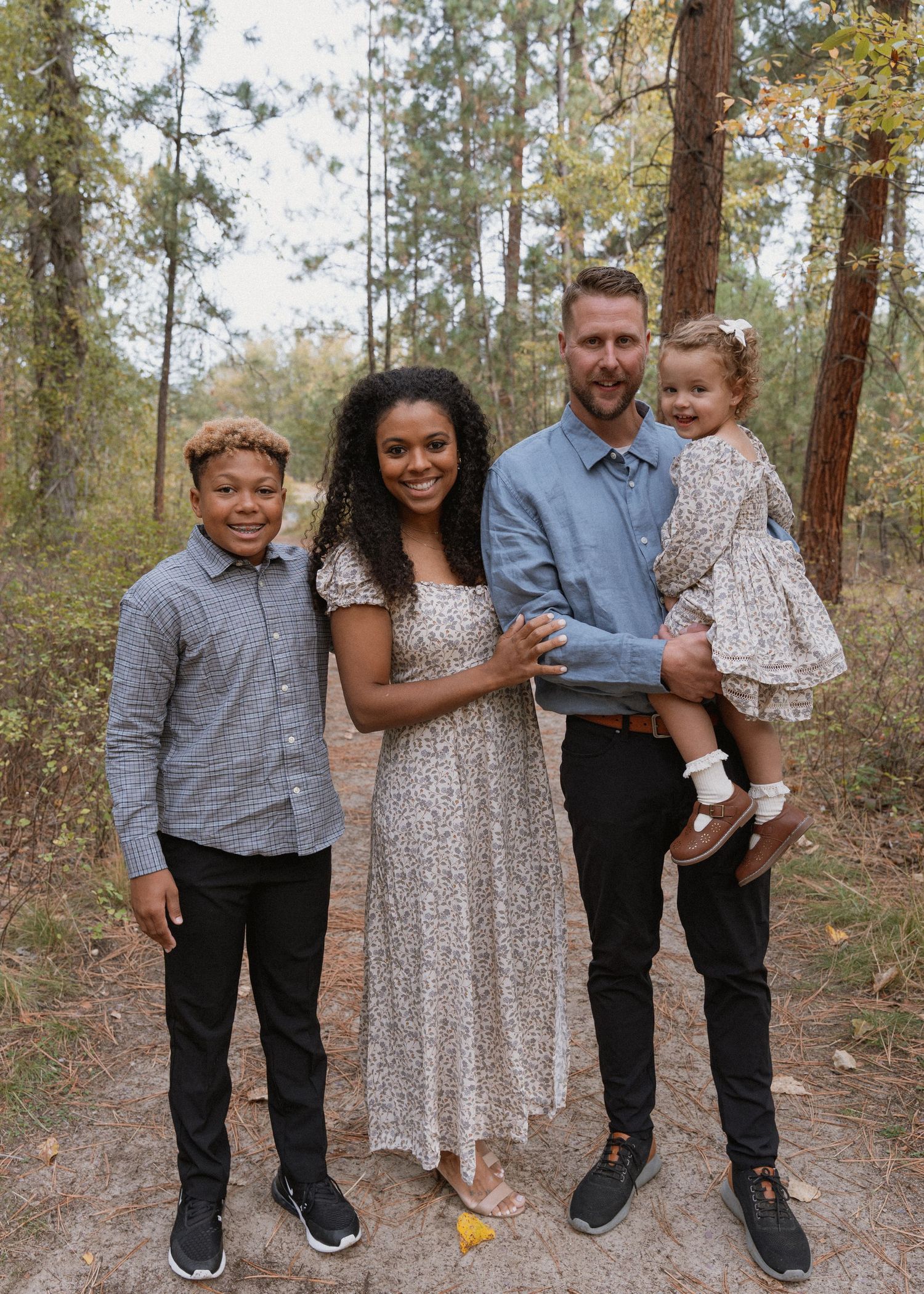 Family of four posing outdoors in a wooded area, wearing coordinated outfits for a professional portrait session.