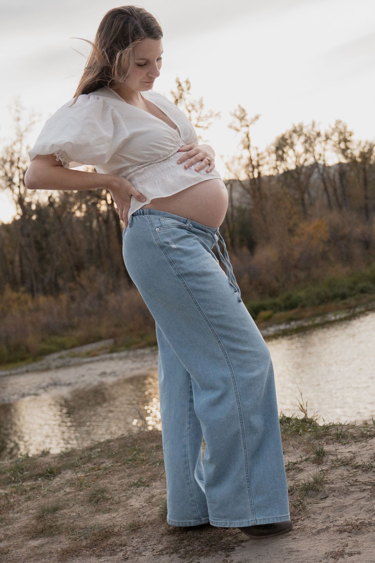 Pregnant woman in a white blouse and wide-leg jeans cradles her bare belly while standing beside a calm river at sunset.