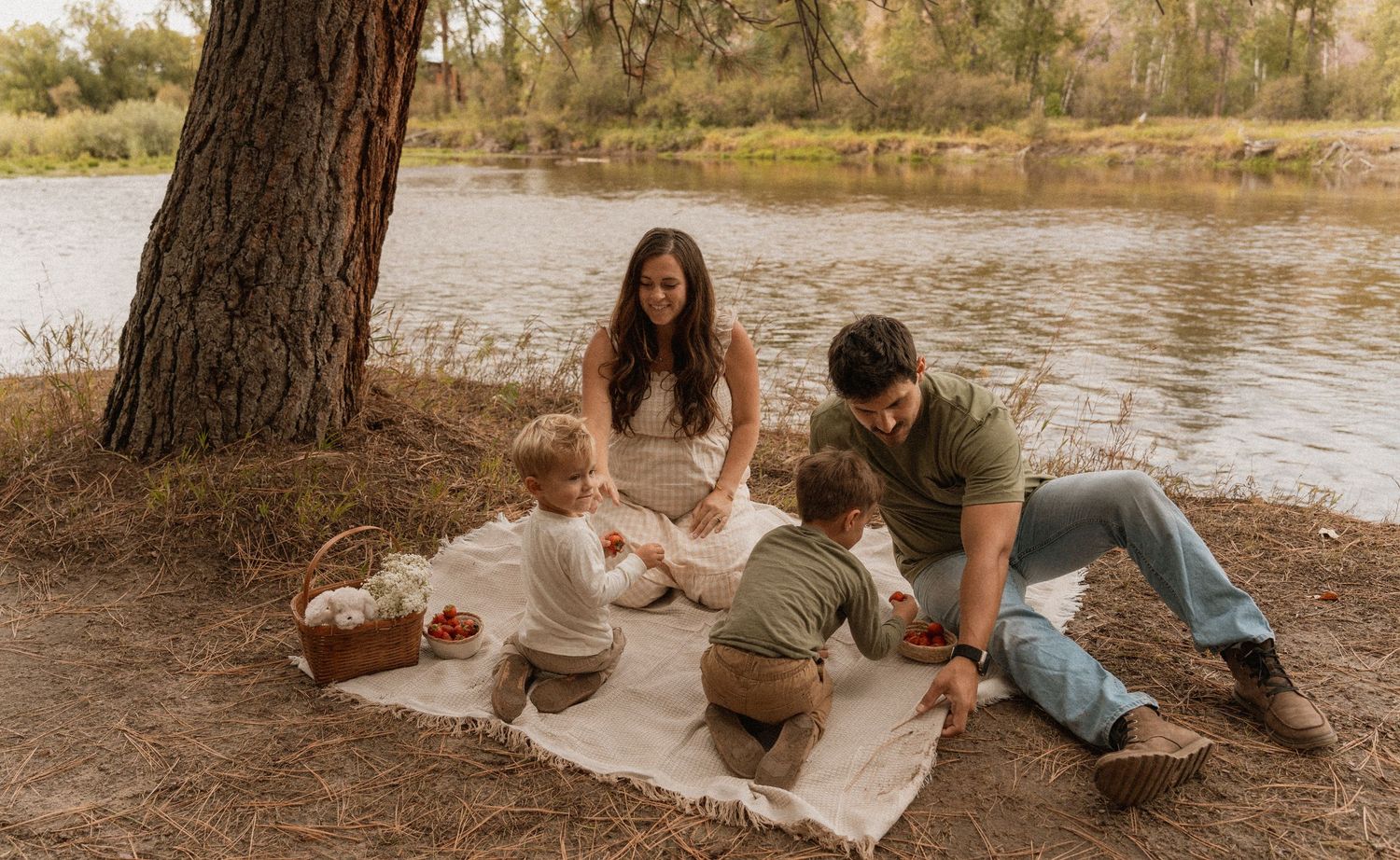 Family enjoying a picnic on a blanket by a river under a large tree on a sunny day.