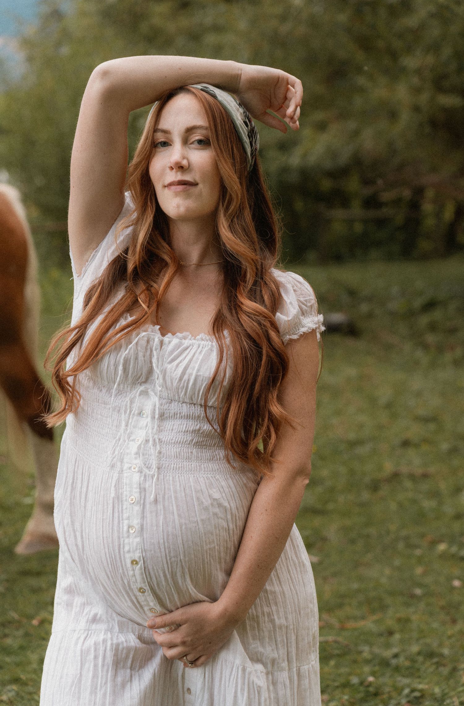 Pregnant woman in white dress with long red hair poses outdoors in green field during maternity photo shoot.
