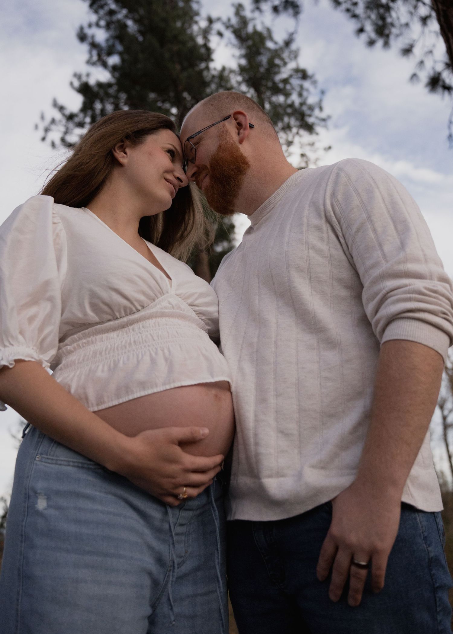 Expectant couple touching foreheads outdoors as the pregnant woman cradles her bare baby bump beneath tall trees and a cloudy sky.