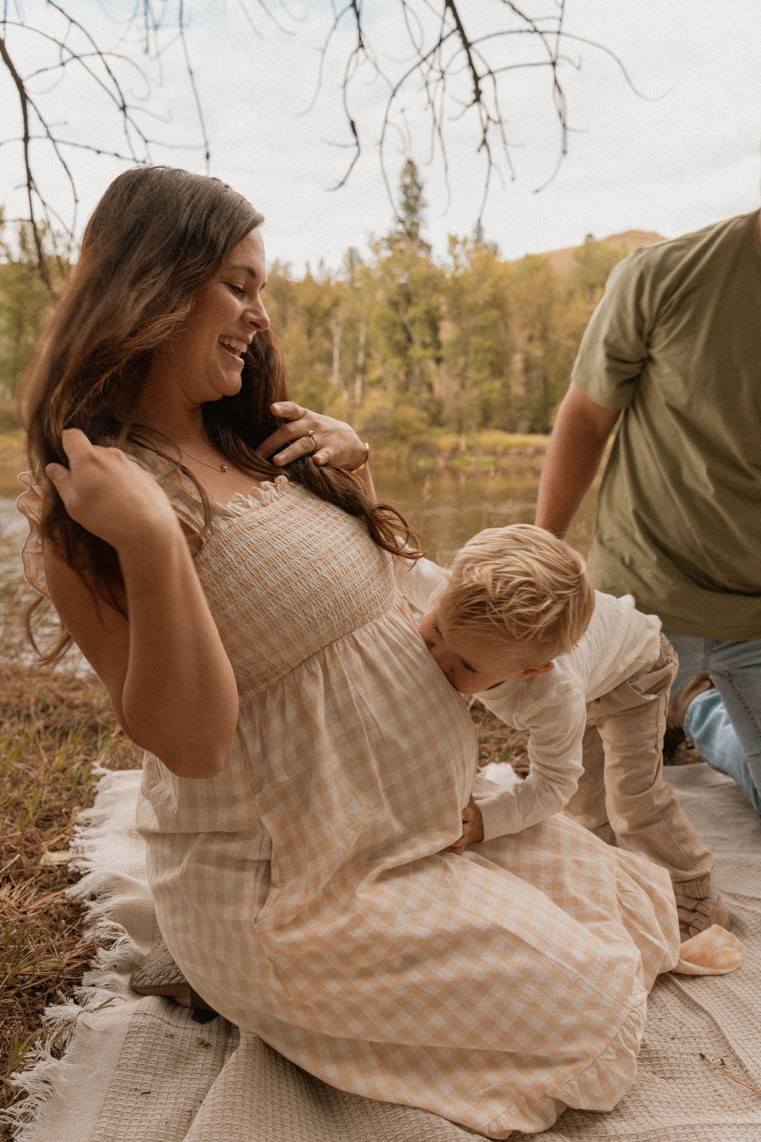 Pregnant woman in beige dress smiling outdoors by lake with toddler and partner during autumn maternity photoshoot.