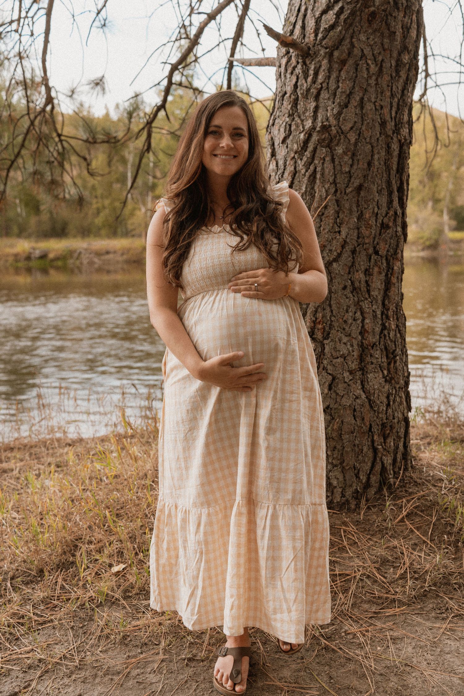 Pregnant woman in a gingham maxi dress cradles her baby bump while standing near a tree by a serene lake.