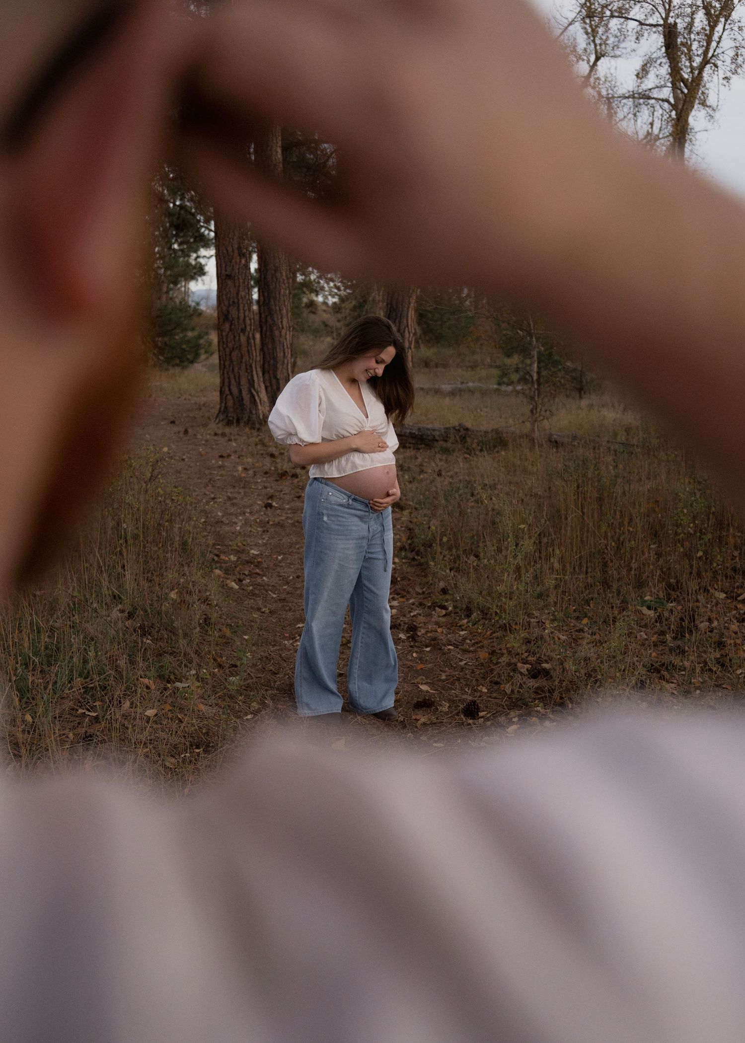 Pregnant woman in a white blouse and blue jeans cradles her baby bump in a sunlit wooded area, framed by blurred hands in the foreground.