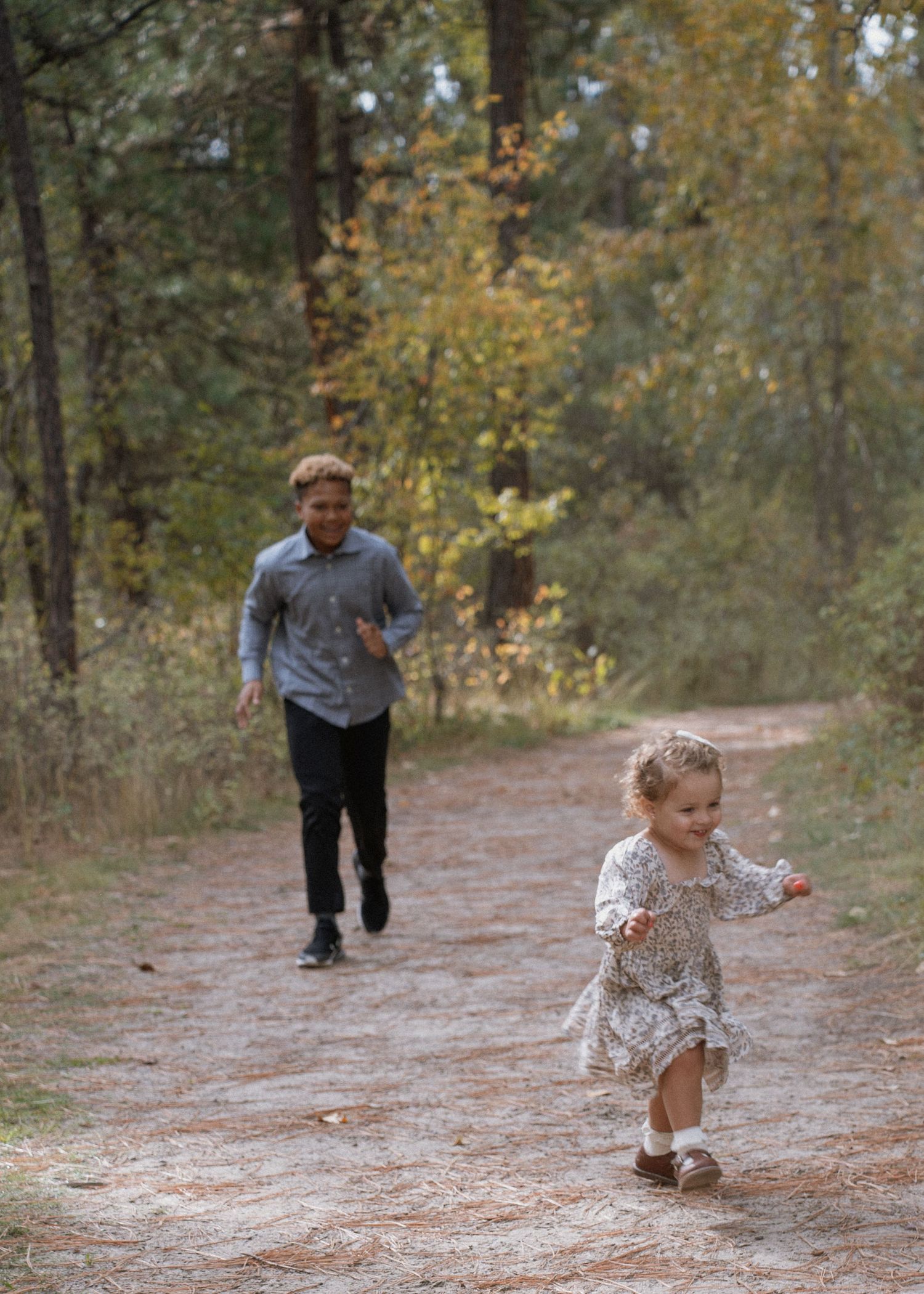 A young girl in a floral dress runs down a forest path as a man chases her during an autumn day.