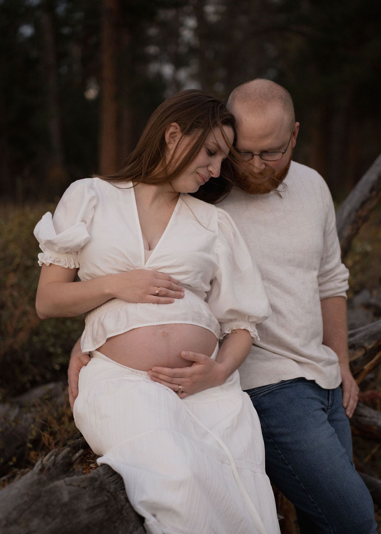 Expecting couple in white and cream outfits embrace outdoors as the woman cradles her bare baby bump during a forest maternity photoshoot.