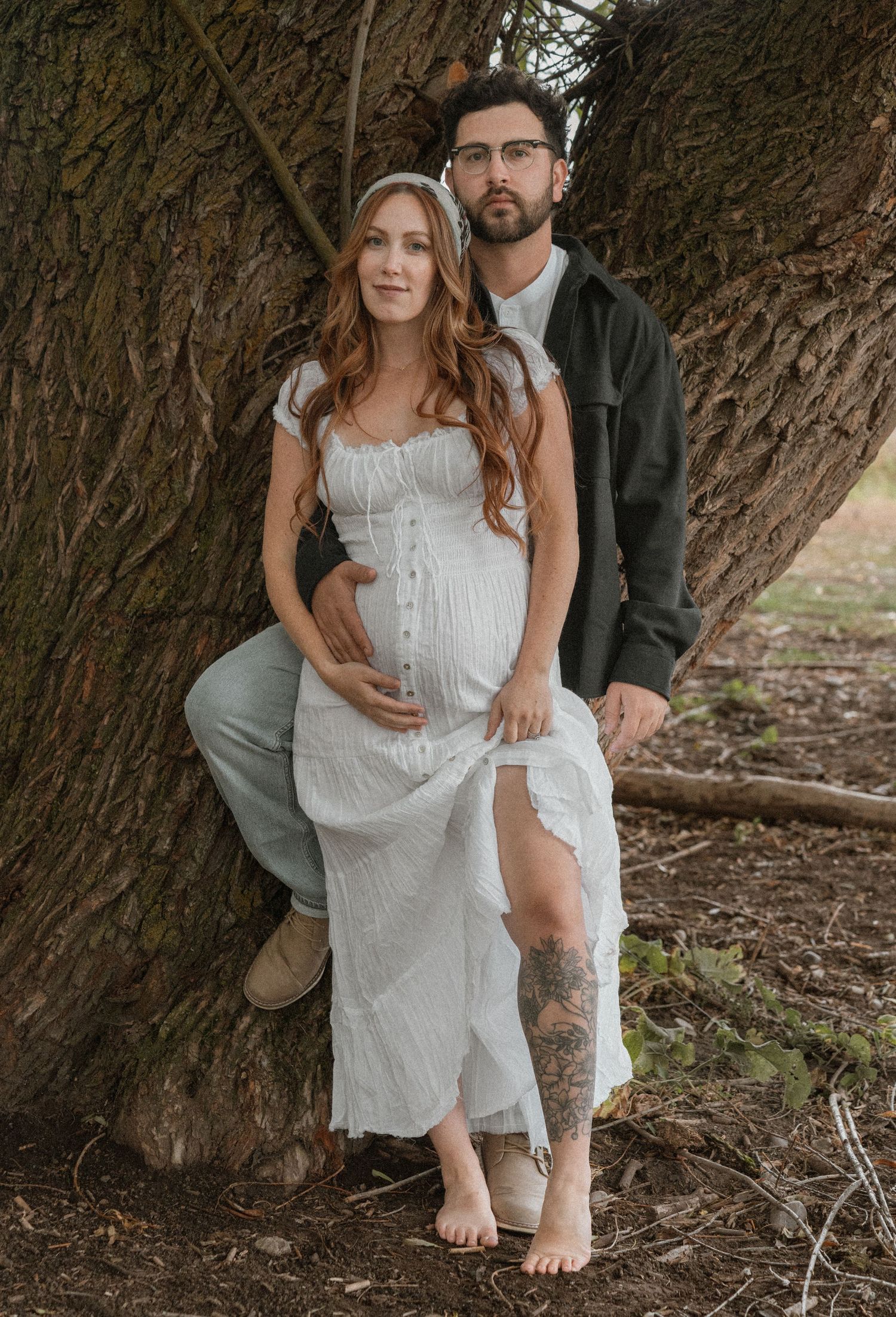 Pregnant woman in white dress poses with bearded man against large tree trunk outdoors for maternity photo.