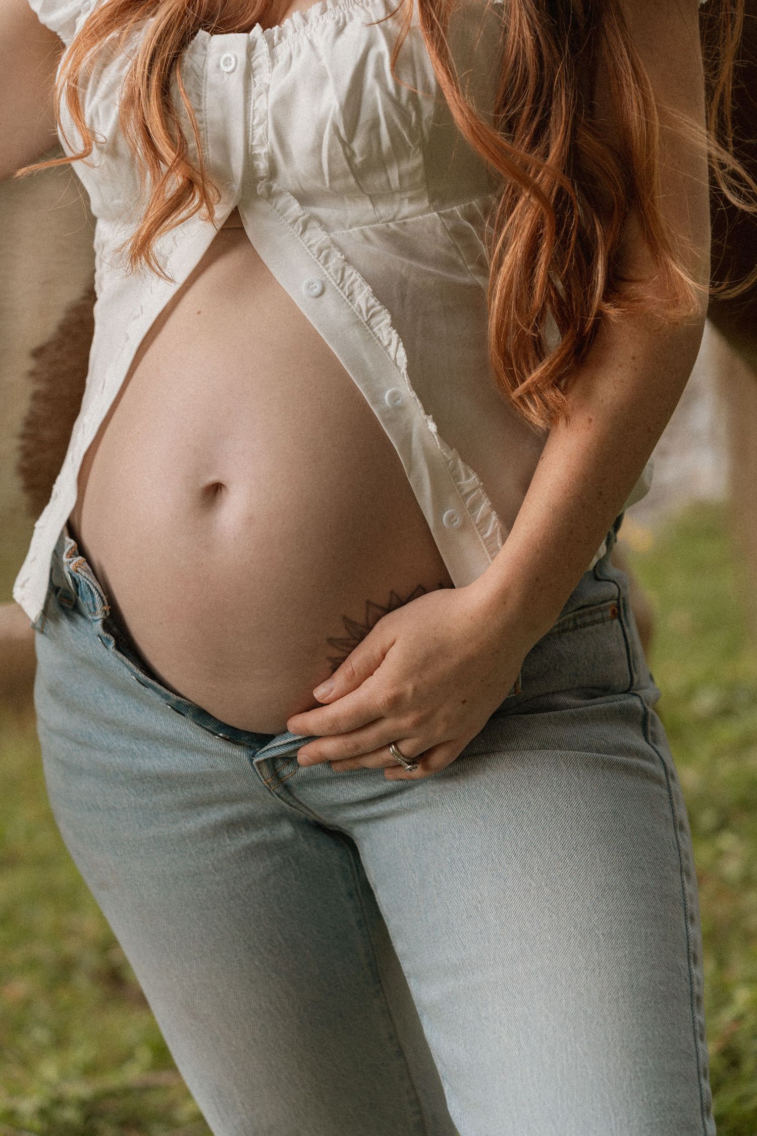 Pregnant woman in white shirt and light jeans cradling her baby bump outdoors.