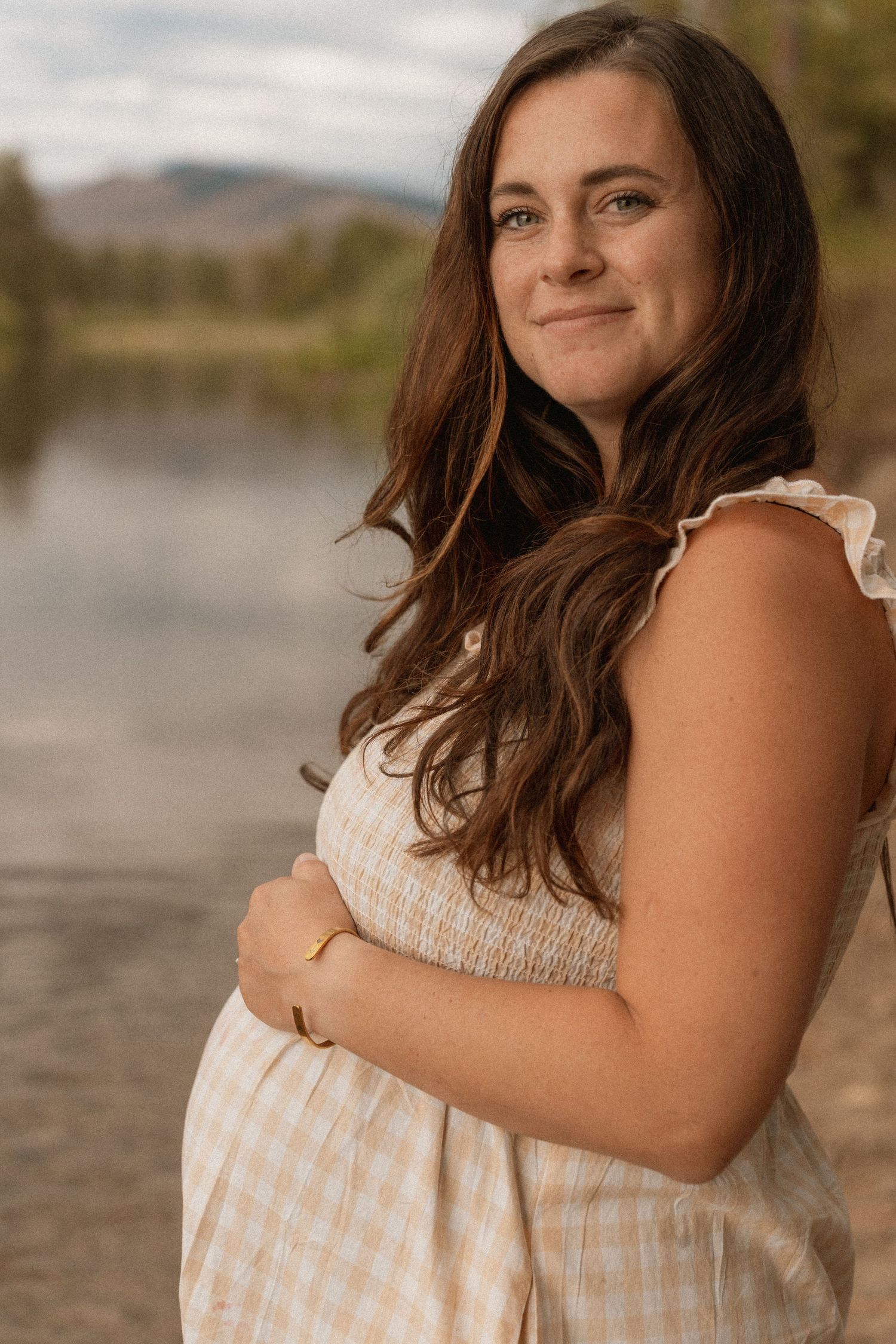 Pregnant woman in gingham dress smiling by a lake with mountains in the background.
