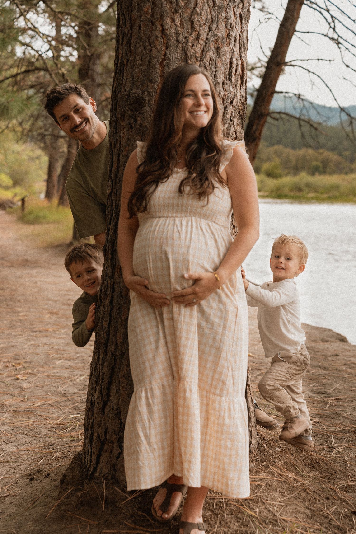 Pregnant woman in white dress poses with family by a tree near a river during an outdoor maternity photo session.