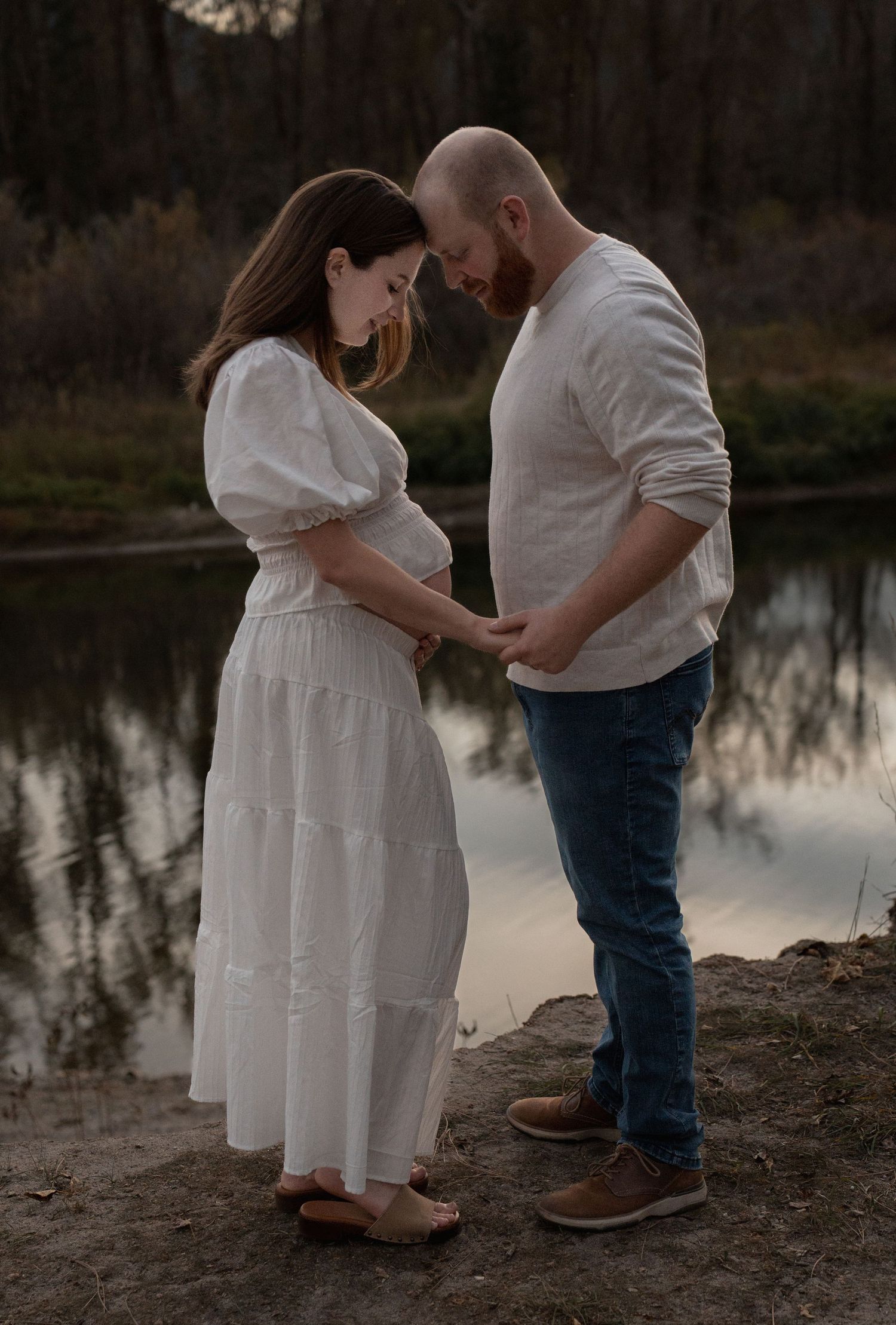 Expecting couple stands forehead to forehead holding hands by a calm reflective river during a maternity photoshoot at dusk.