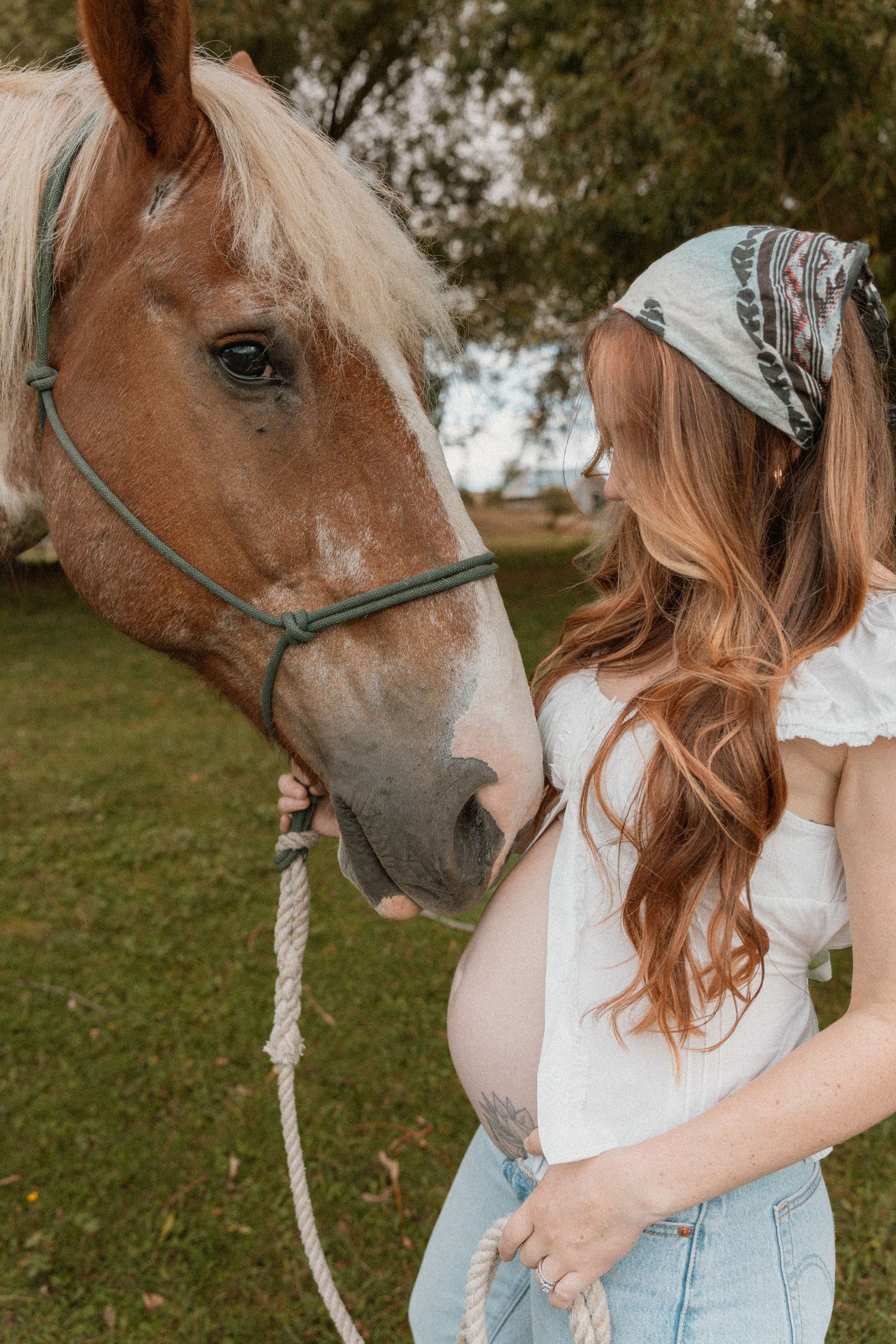Pregnant woman in white top and jeans holds rope lead of brown horse outdoors on green grass.