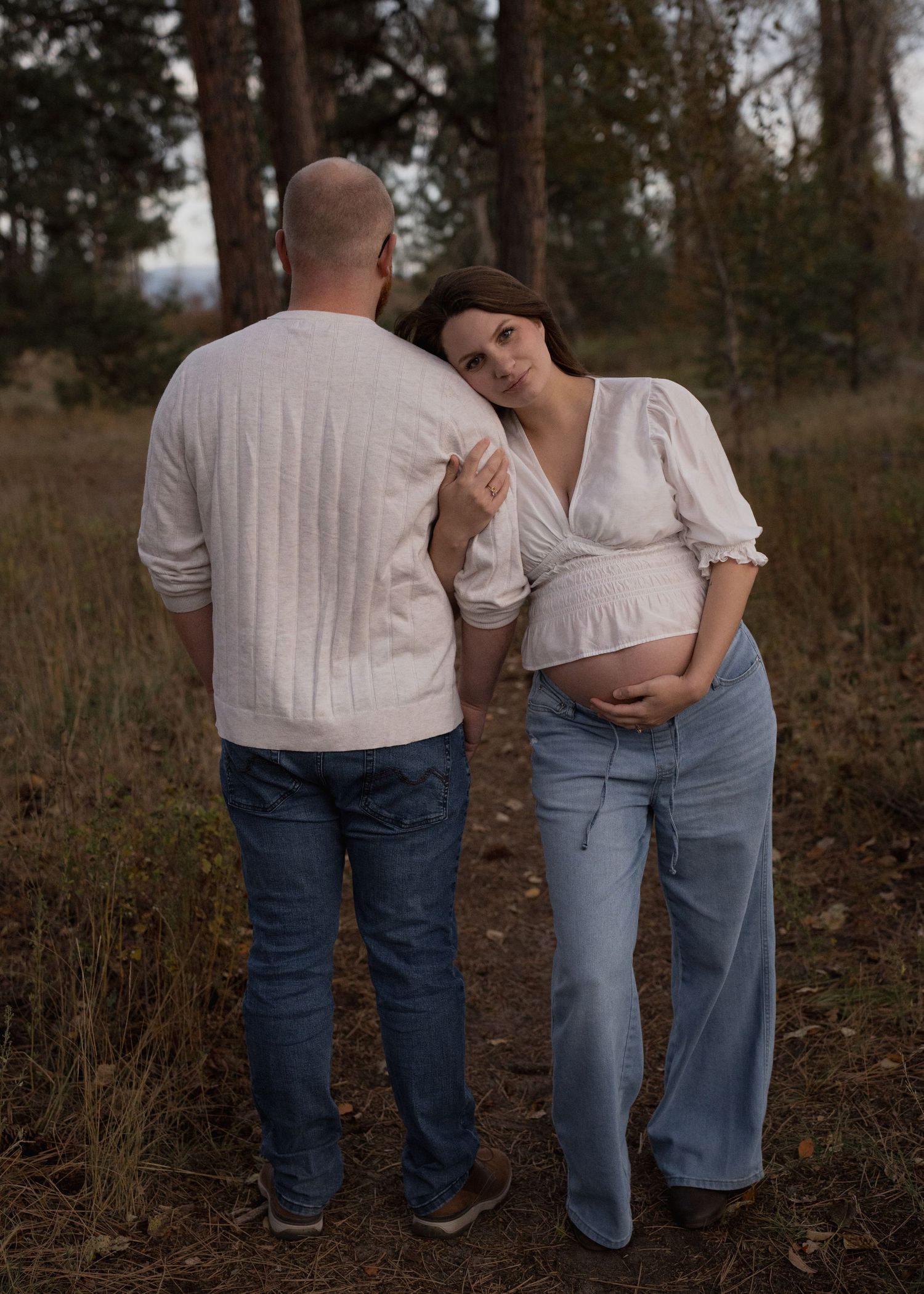 Expecting couple in a woodland maternity photo, the pregnant woman in a white blouse and jeans resting her head on her partner's shoulder.