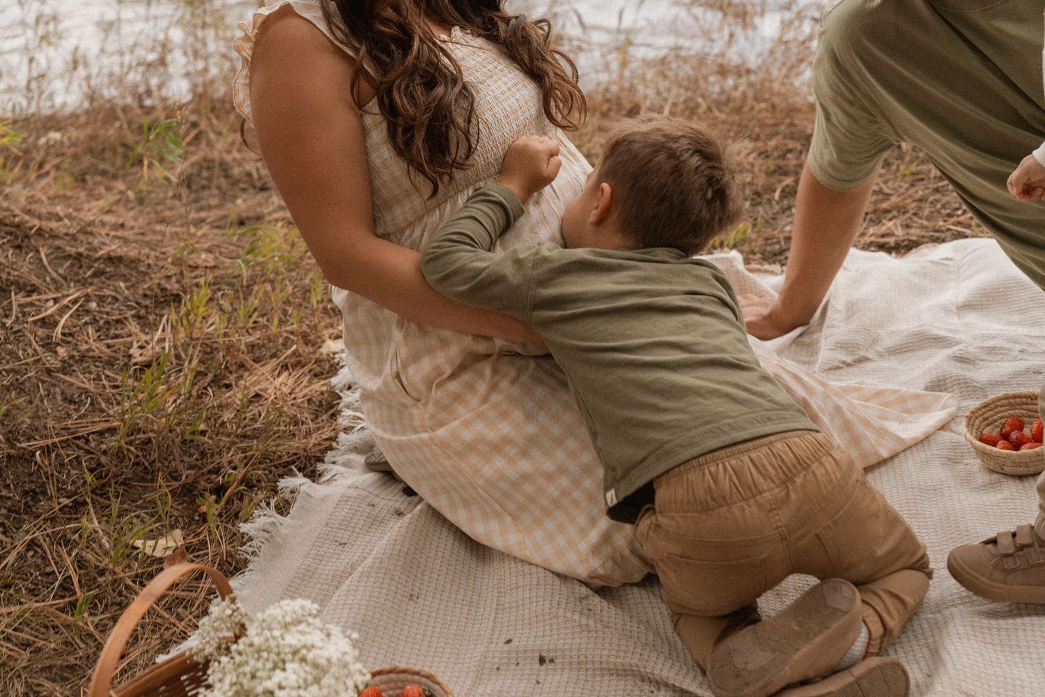 A toddler in green shirt kneels on a white blanket outdoors, reaching toward a woman in a plaid dress.