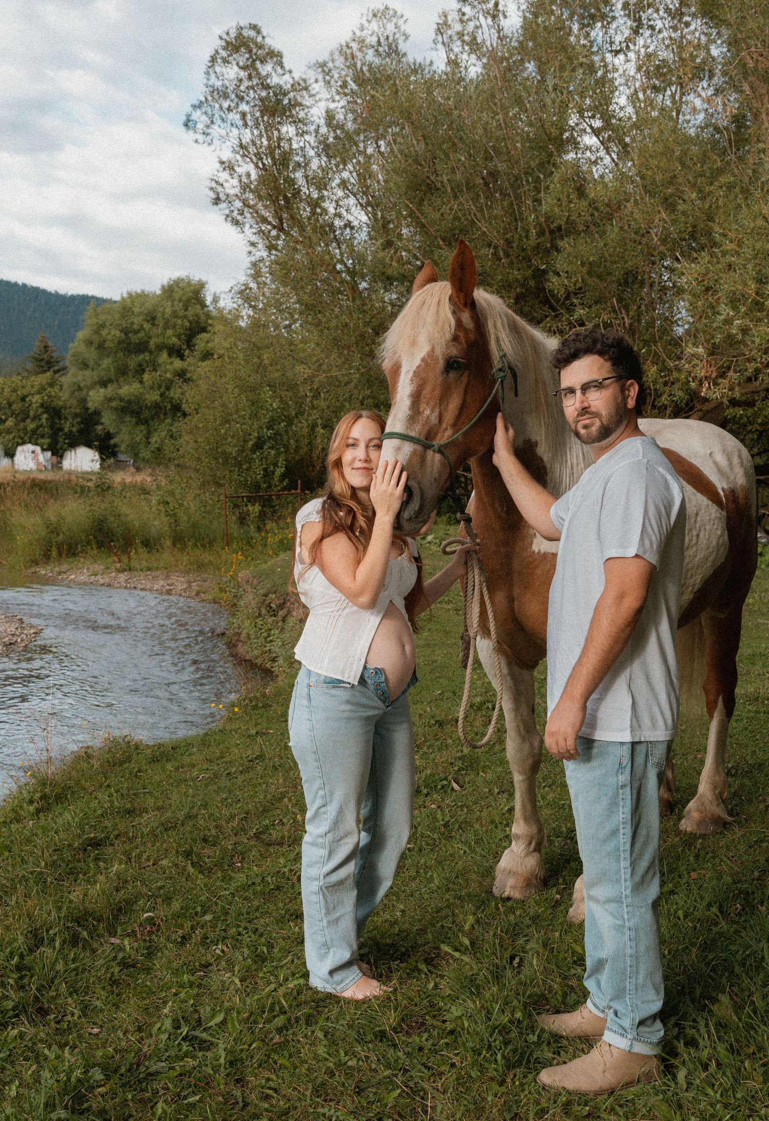 Pregnant couple standing with a brown and white horse by a river surrounded by lush green trees.