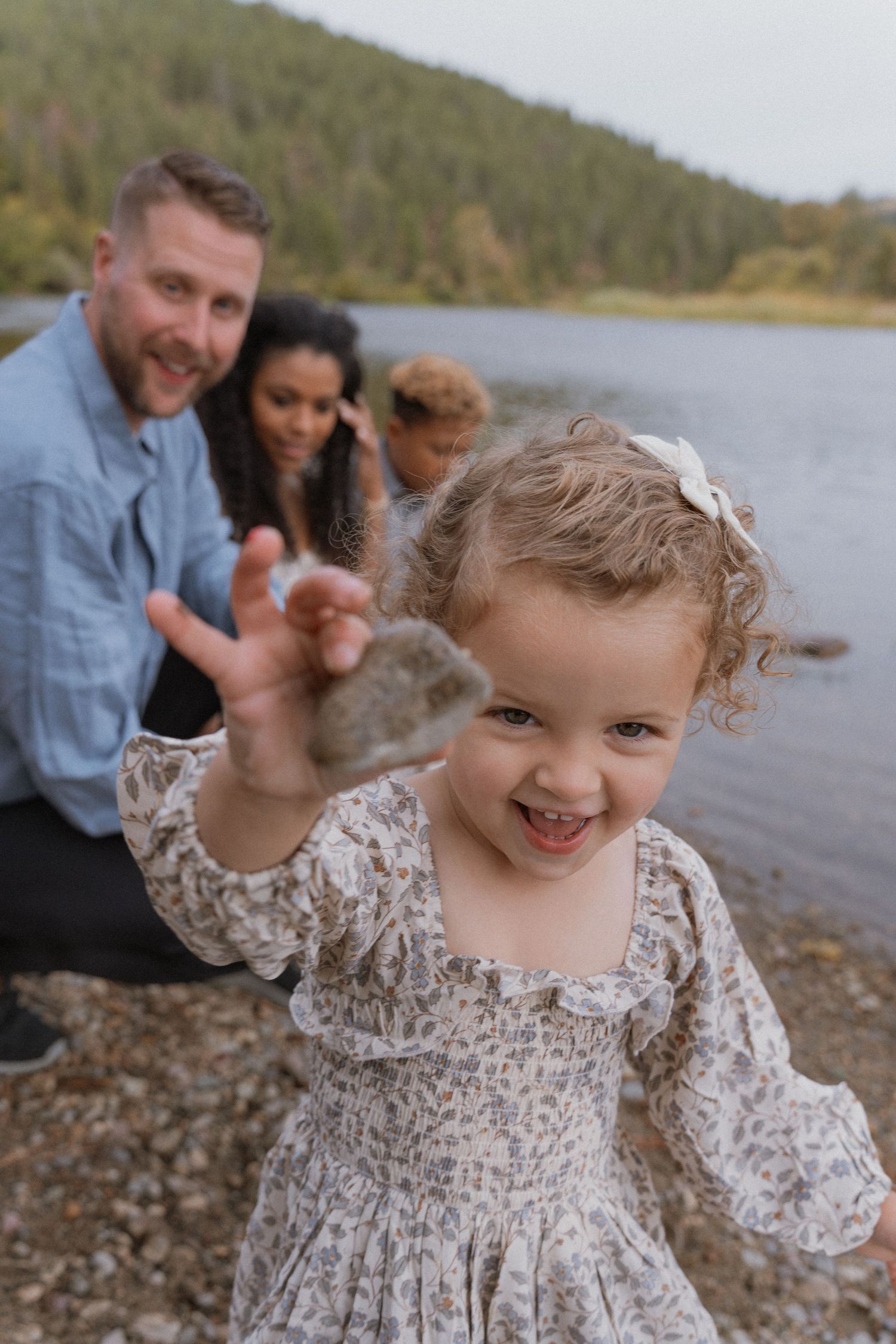 A young curly-haired girl in a floral dress holds a rock near a lake as her family smiles behind her.
