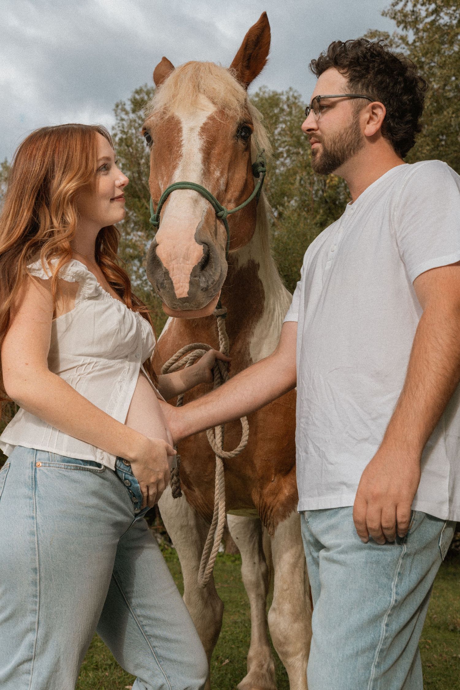 Pregnant woman with red hair and man in glasses stand with a brown and white horse outdoors.