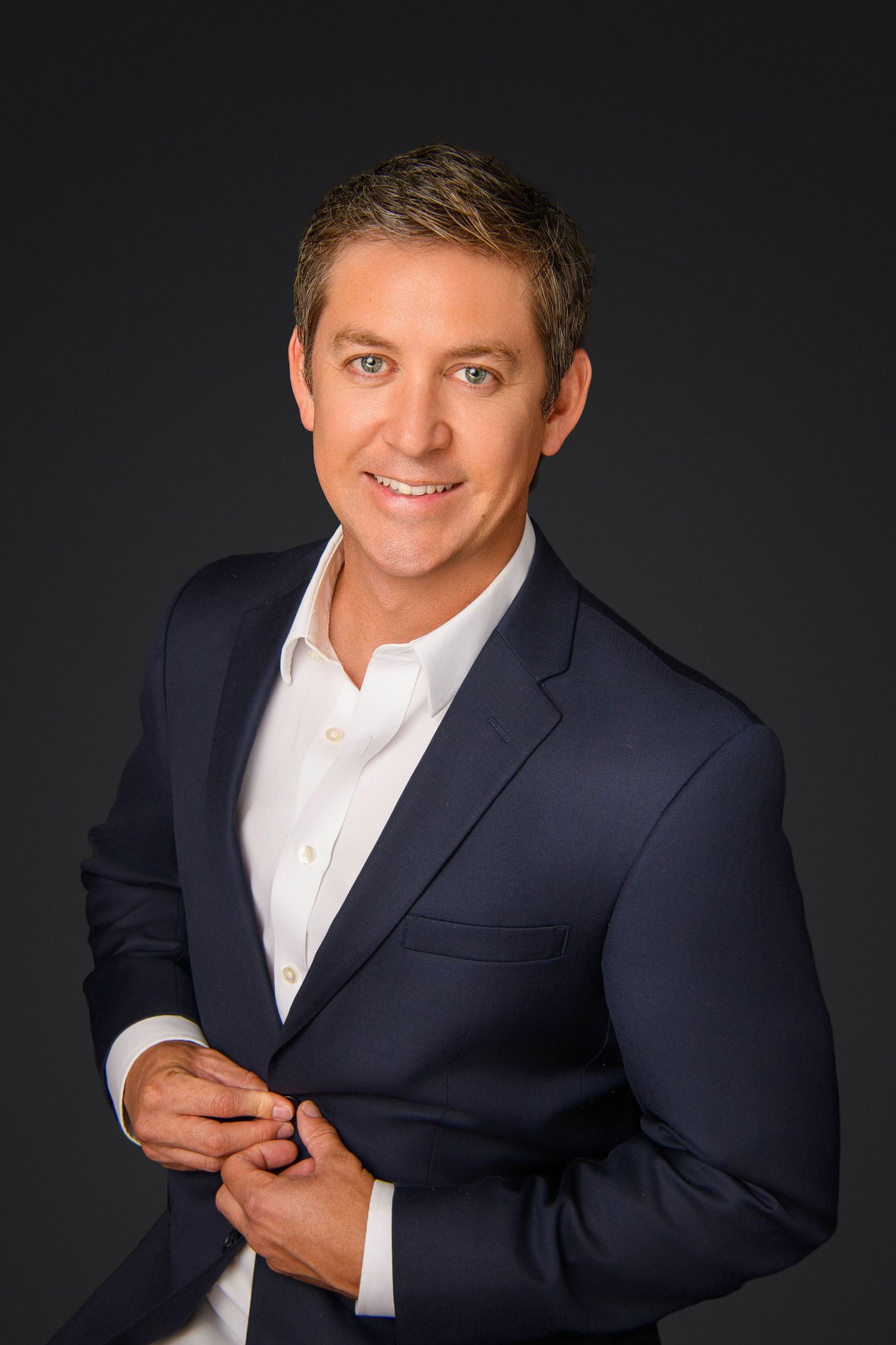 Professional man in navy blazer and white shirt smiling confidently against dark gray background.