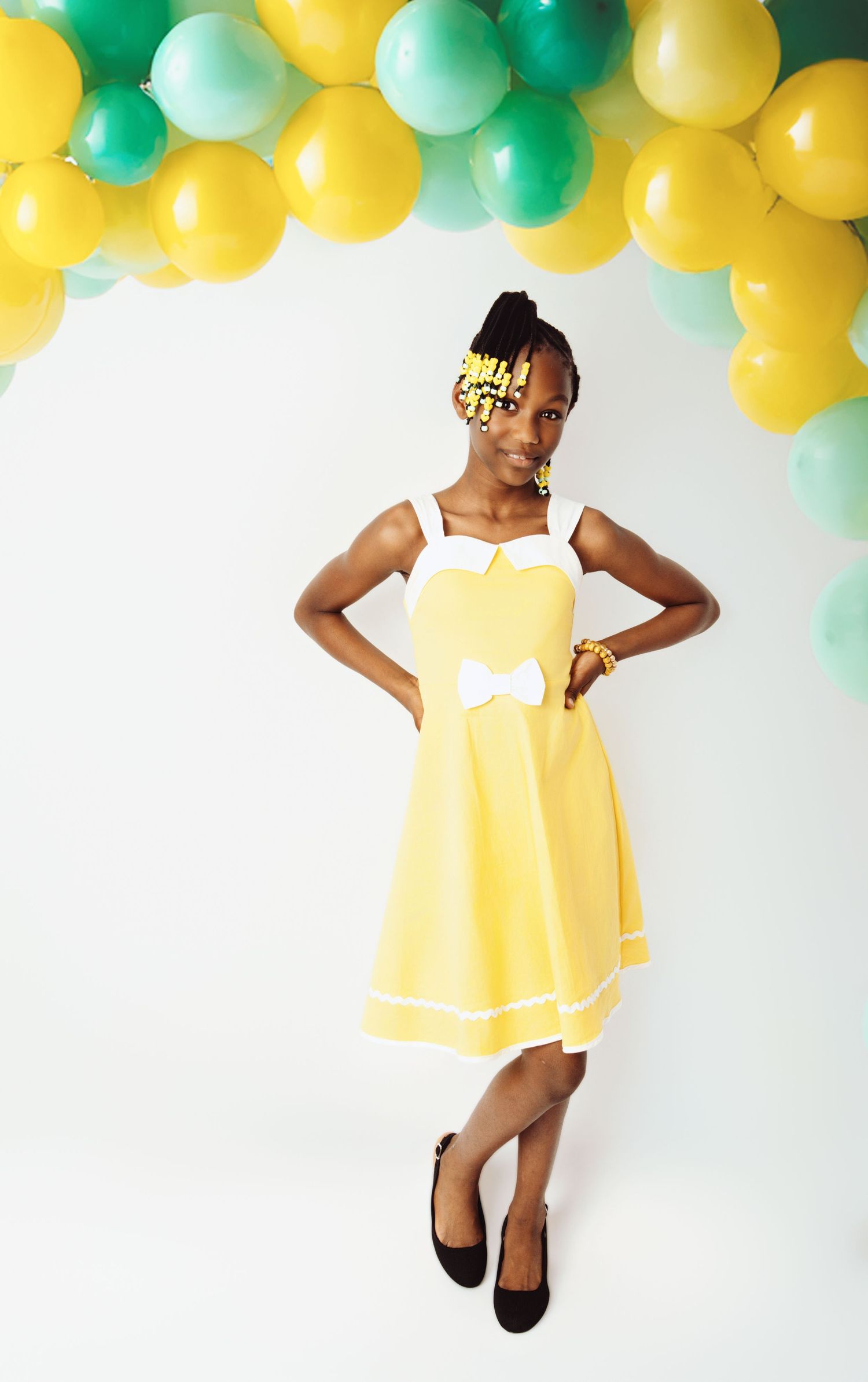 Child in yellow dress posing under festive yellow and green balloon arch.