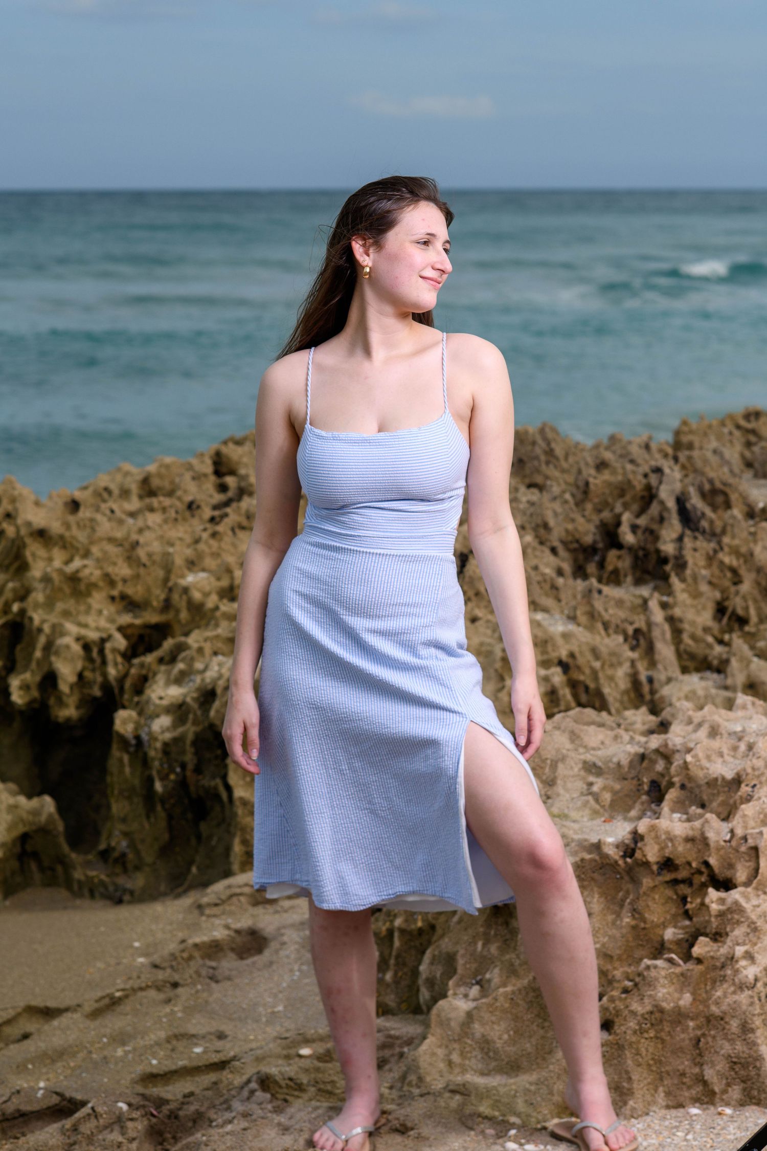 A casual beach photoshoot featuring a light blue tank top against coastal rocks and ocean waves.