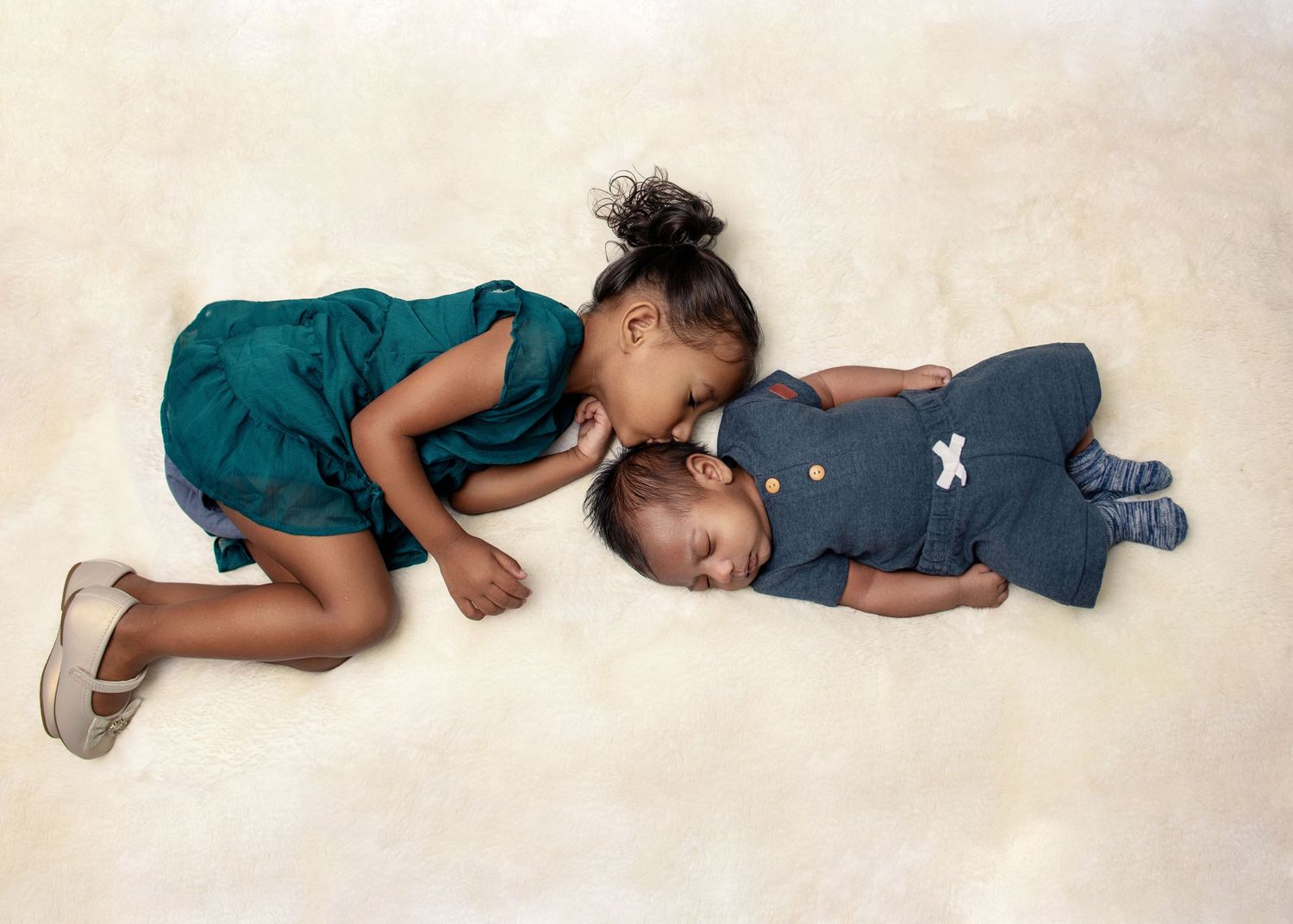 Two children in green and blue outfits lying on cream colored carpet.