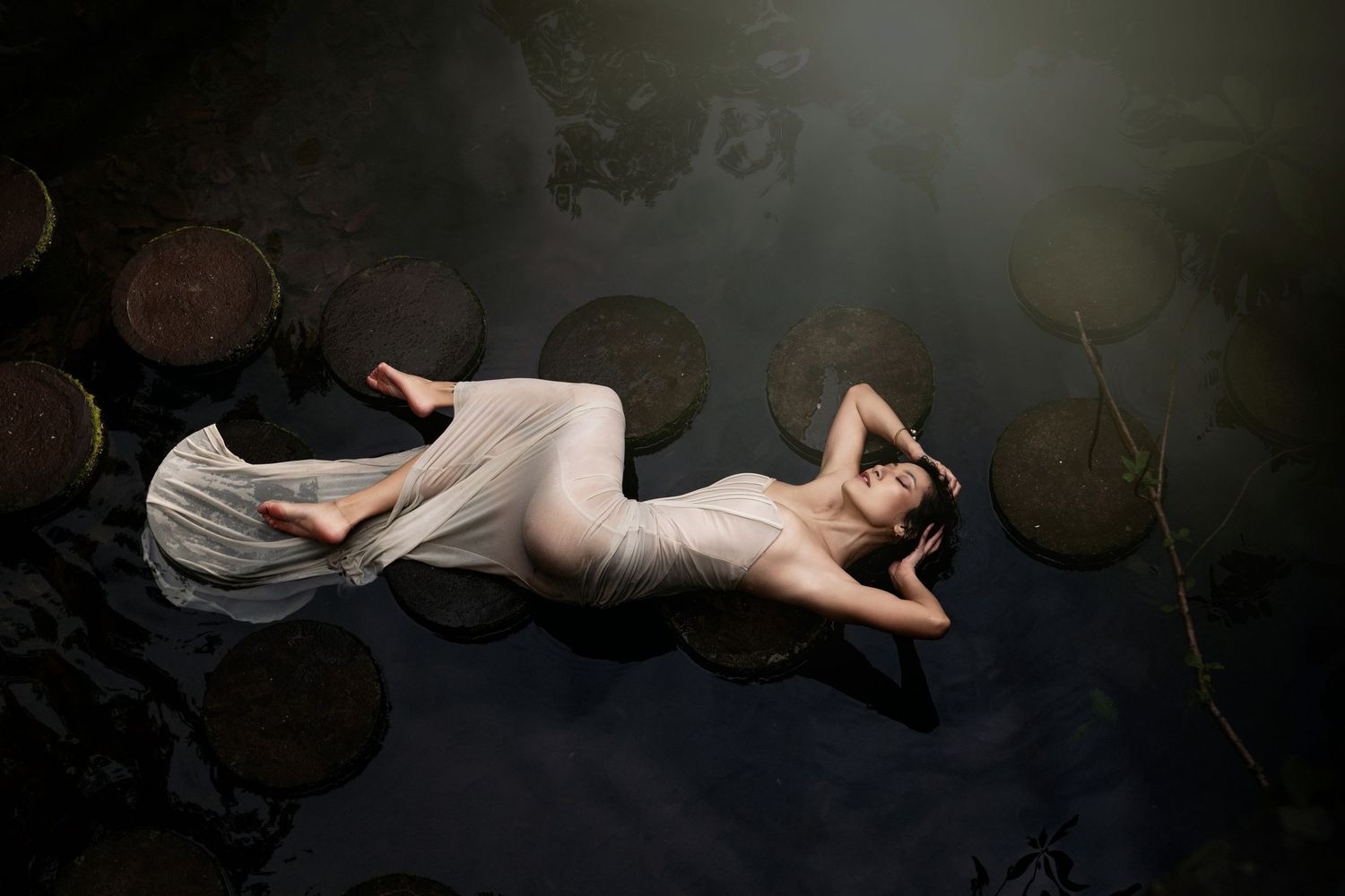 Artistic portrait of a figure in white fabric lying on dark reflective surface near lily pads.