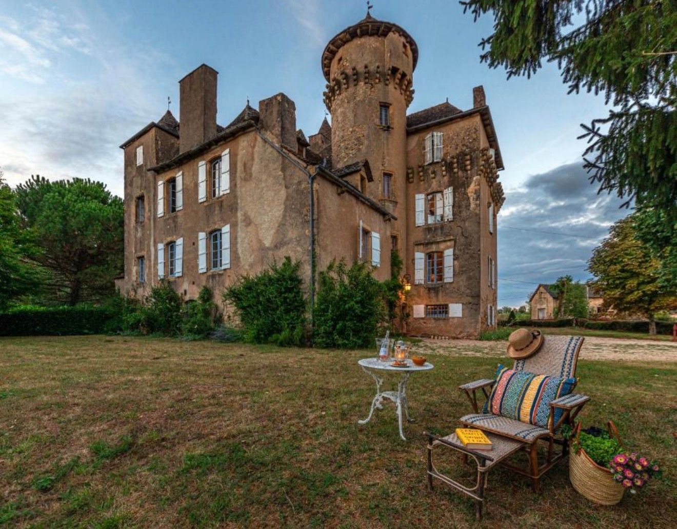 Historic French château with round tower, brick façade, and garden seating area under a dramatic evening sky.