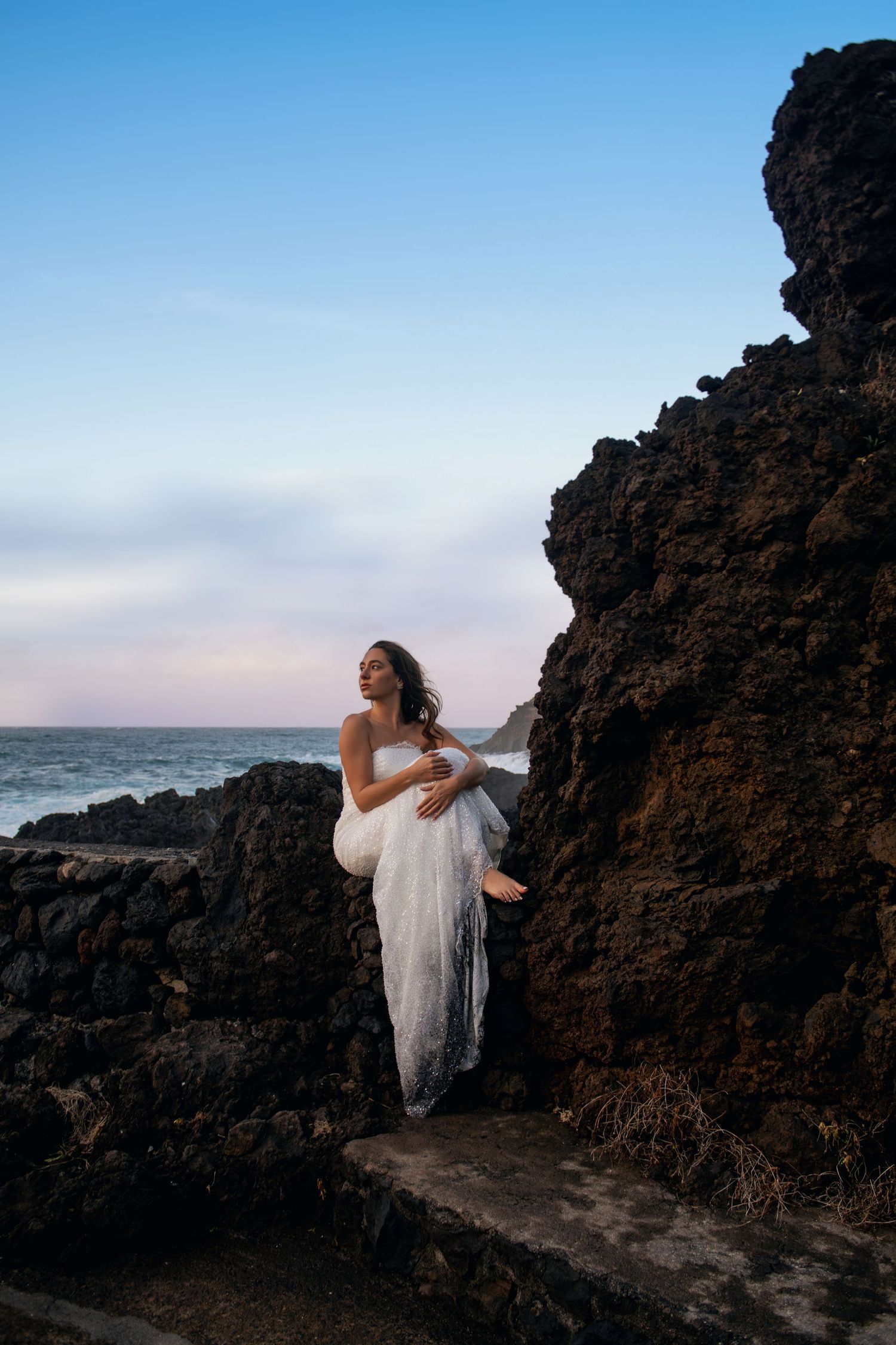 Figure in white dress climbing on volcanic rocks near ocean at sunset.
